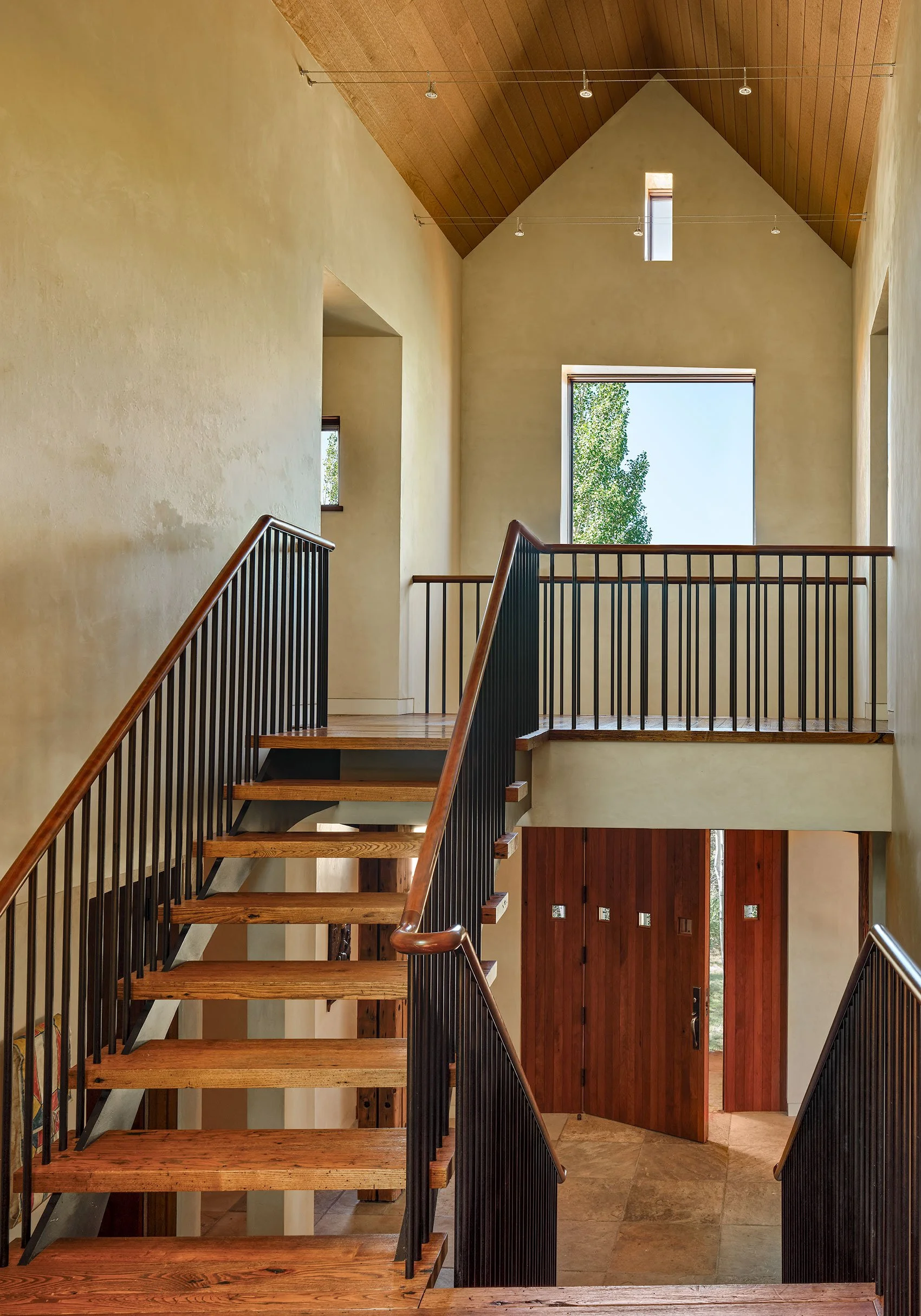 Interior view of a staircase with wooden steps and black metal railings, leading to a second floor with a large window showing trees outside, a wood-paneled ceiling, and a wooden door at the bottom of the stairs.