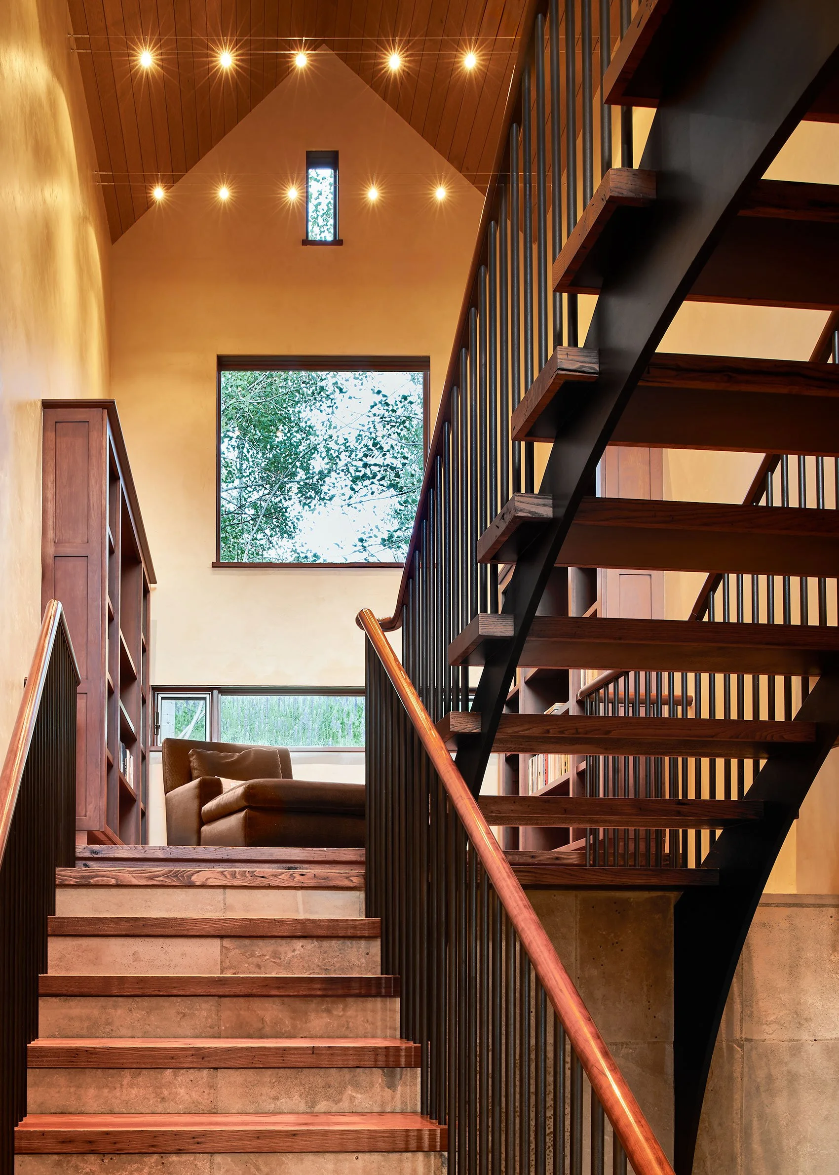 Interior view of a stairway with wooden steps, black metal railings, and a brown wooden handrail, with large windows showing trees outside and warm lighting on the ceiling.