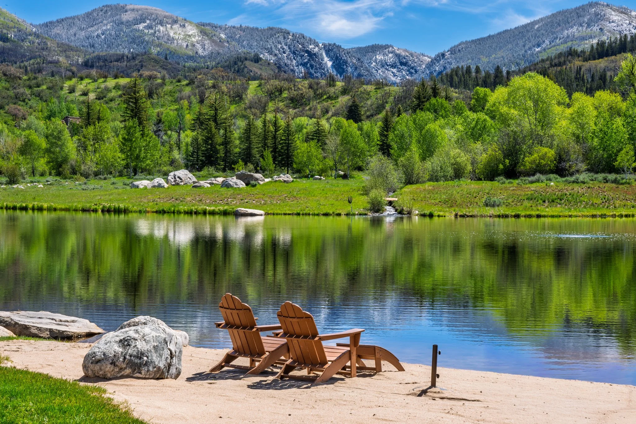 A peaceful lakeside scene with two wooden lounge chairs on a sandy beach, large rocks, and lush green trees, with a calm lake reflecting the surrounding scenery and snow-capped mountains in the background under a partly cloudy sky.