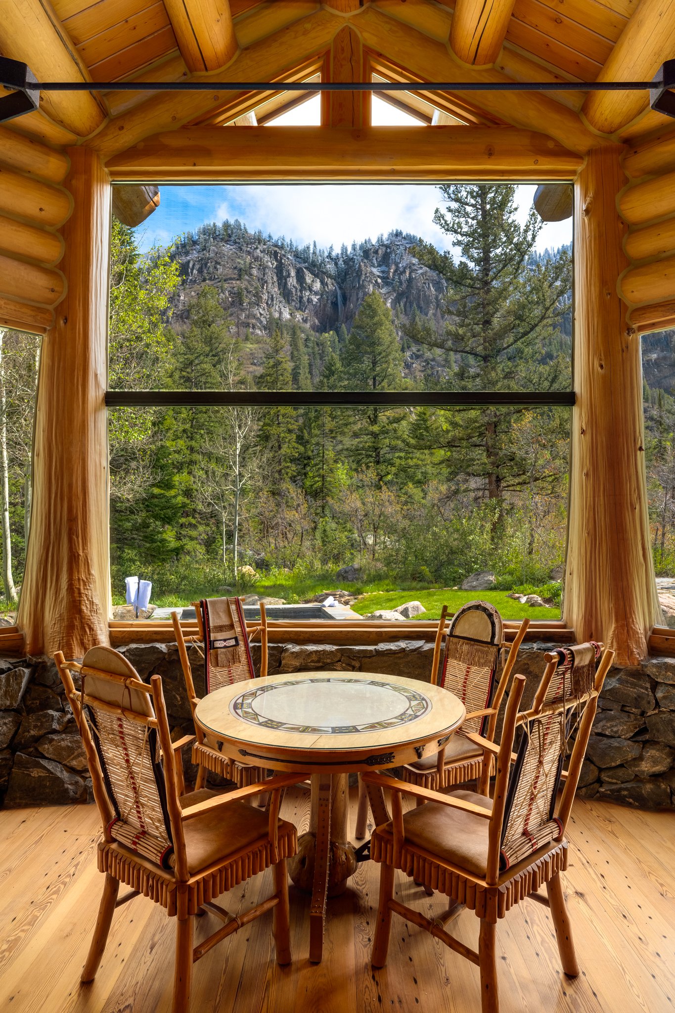 View from inside a log cabin, looking out a large window at a forest and mountains. Sitting at a wooden table with chairs around it inside the cabin.