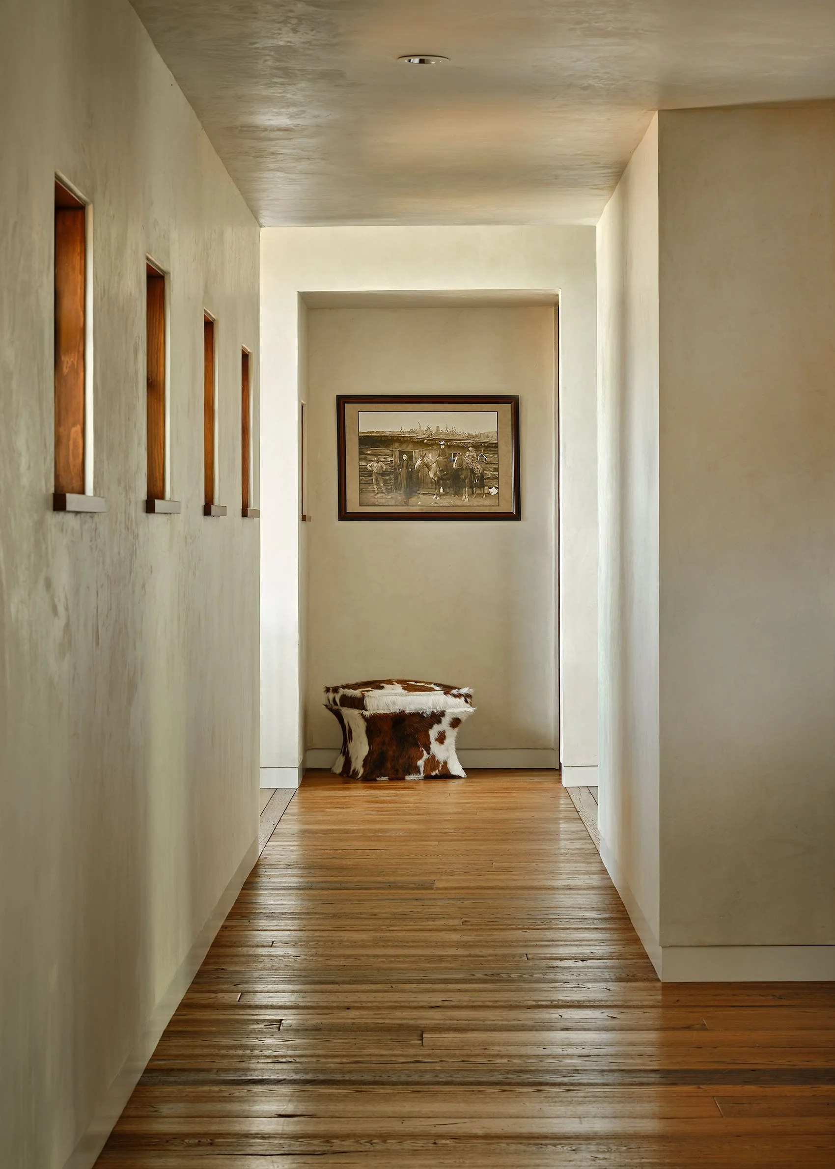 A hallway with hardwood flooring and cream-colored walls. There are four narrow vertical windows on the left wall and a framed black-and-white photograph at the end of the hallway. A cowhide upholstered stool is positioned in front of the photograph.