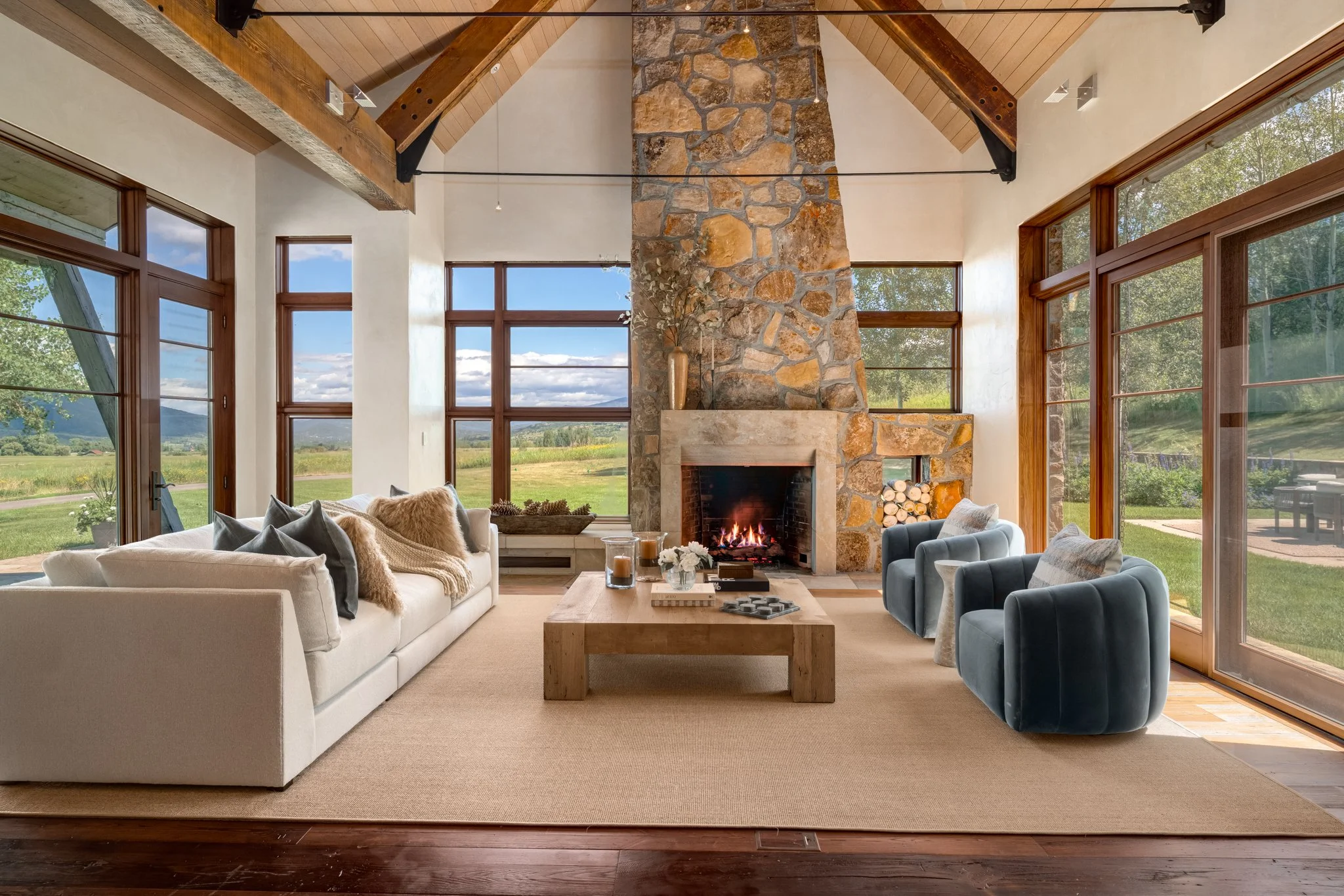 Living room with large windows showing a scenic outdoor landscape, stone fireplace with fire burning, various sofas and armchairs, wooden ceiling beams.