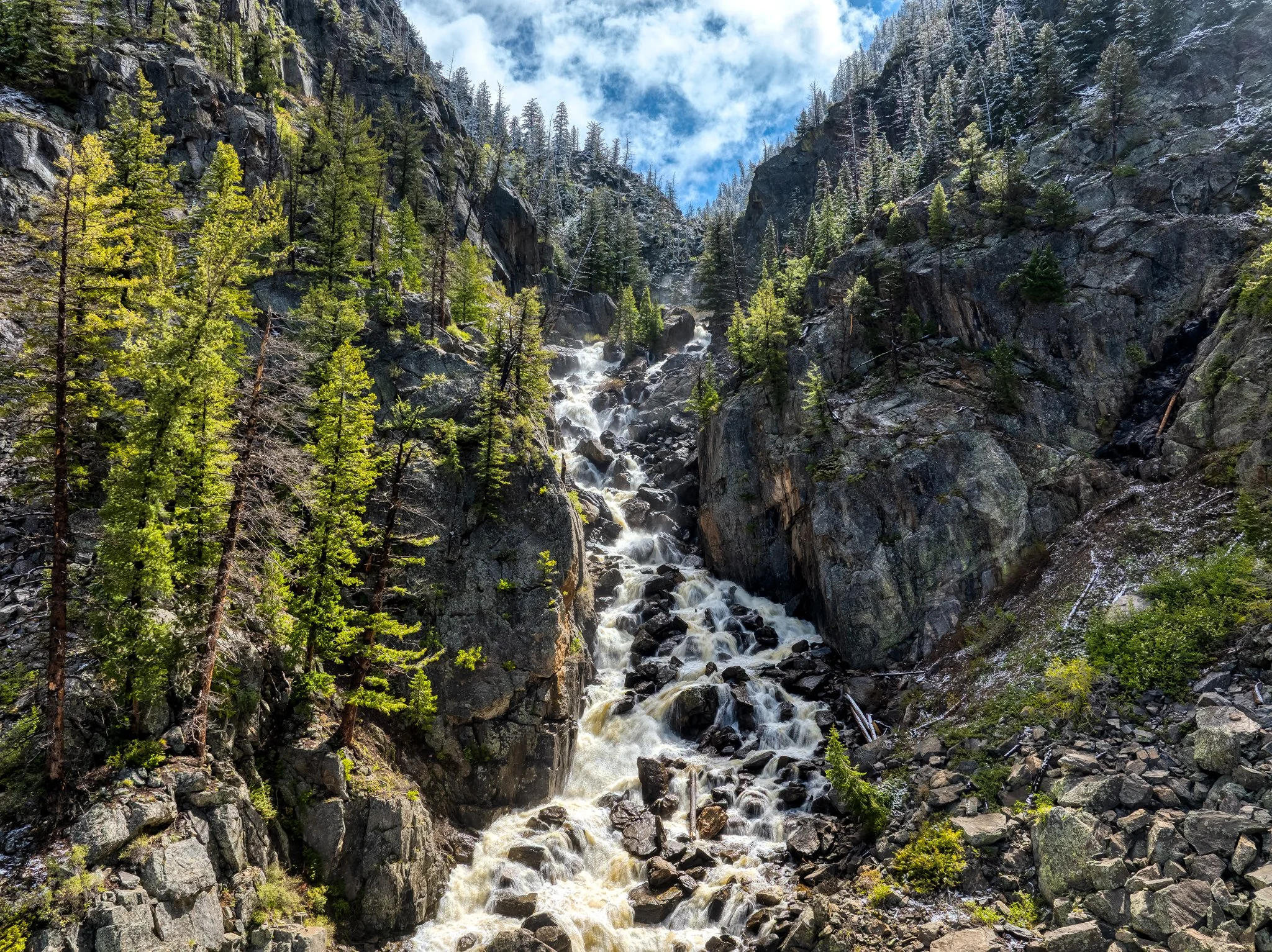 A mountain landscape with a waterfall flowing down rocky cliffs flanked by green pine trees, under a partly cloudy sky.
