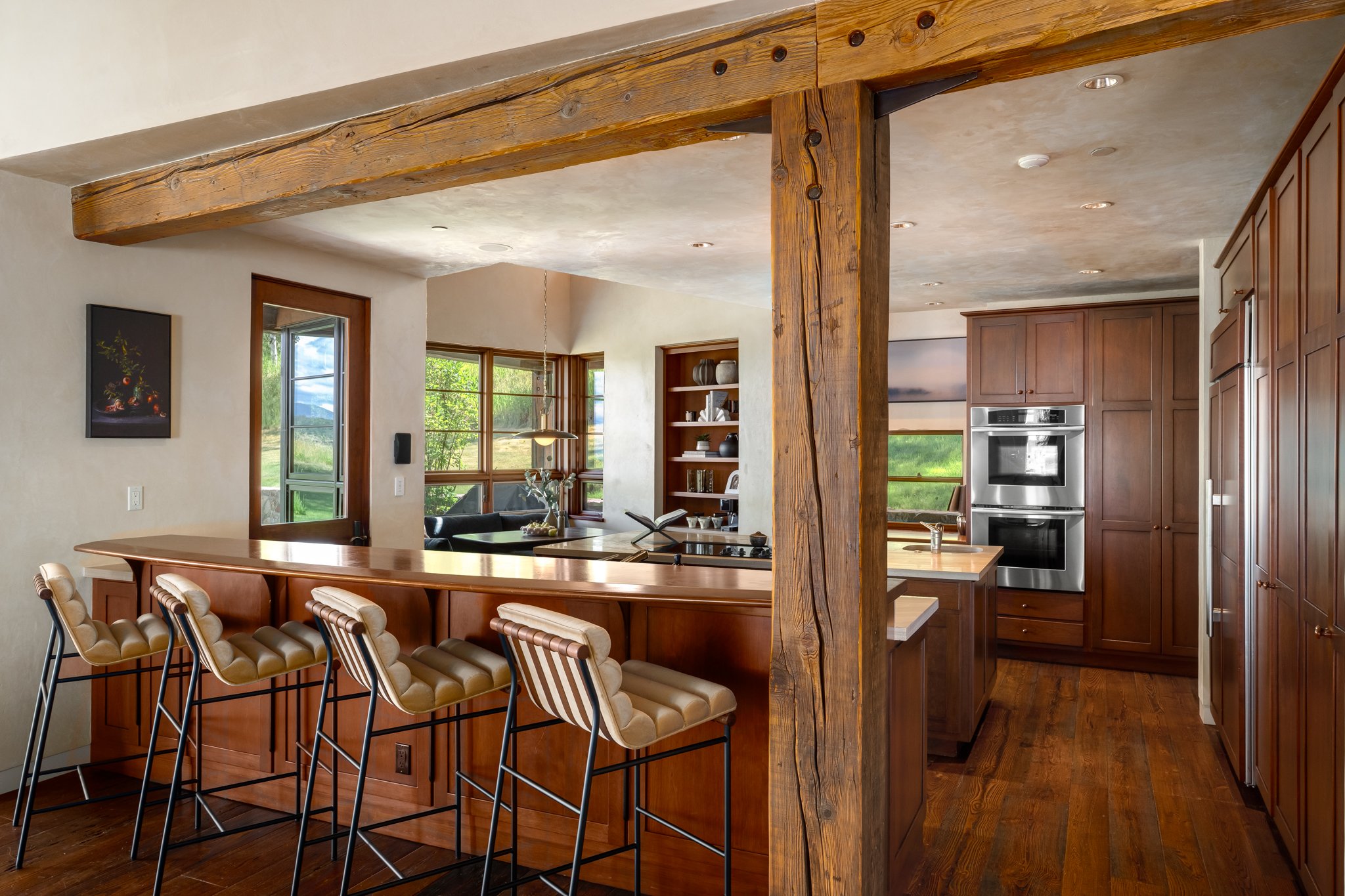 Open-concept kitchen with a wooden beam ceiling, large windows, and a breakfast bar with beige cushioned stools.