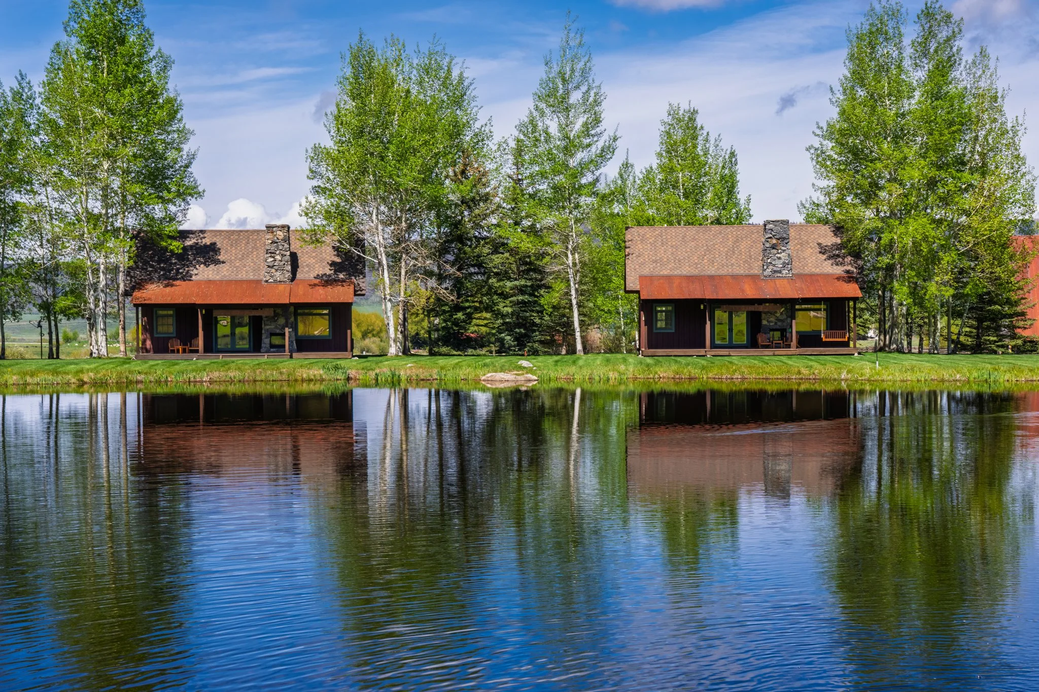 Two houses with dark wood siding and red metal roofs are situated on a grassy area beside a body of water, surrounded by tall green trees under a partly cloudy blue sky.