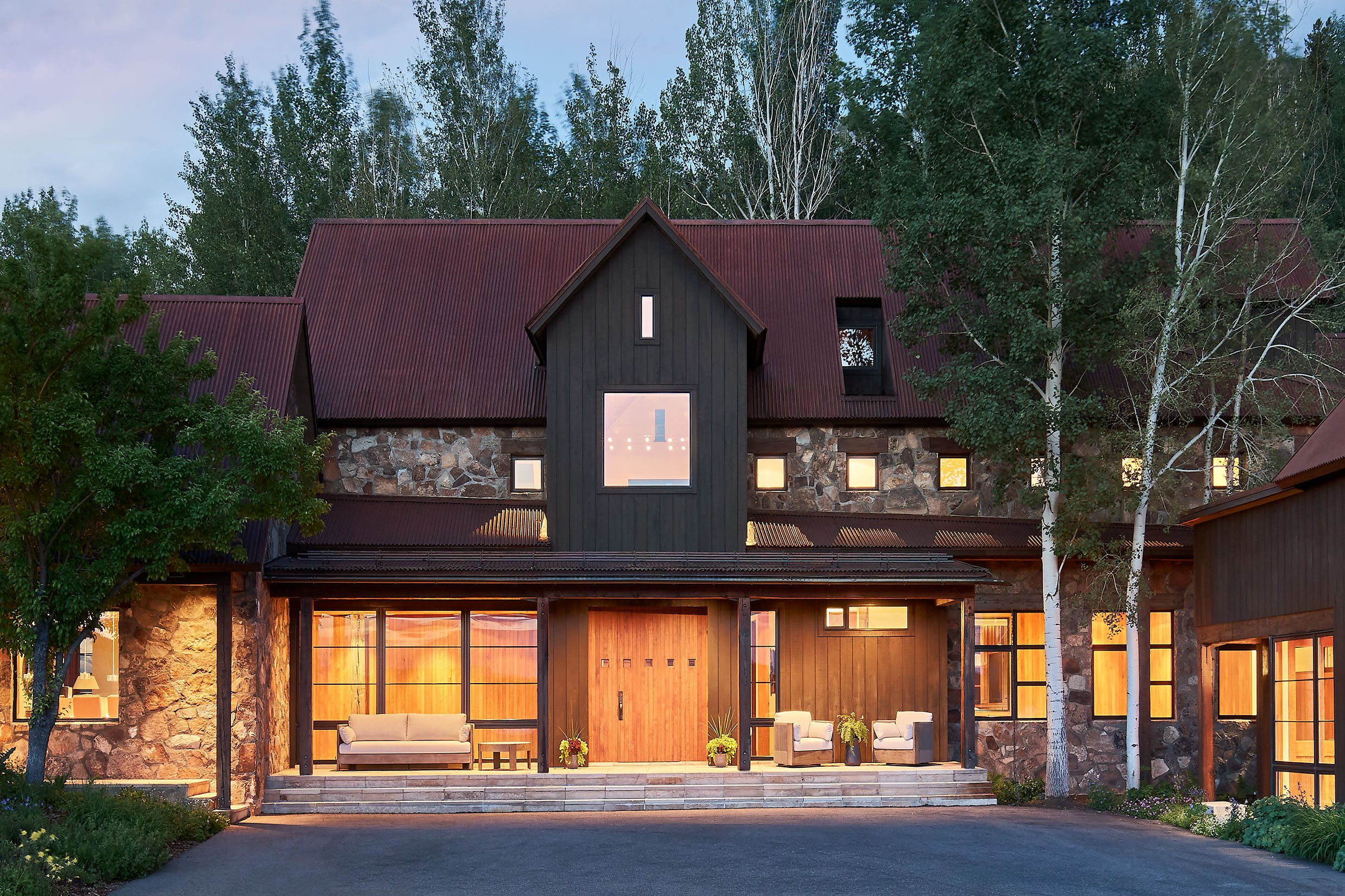Modern house with large windows, stone and wood exterior, red metal roof, surrounded by trees and a lawn, illuminated from inside during evening.
