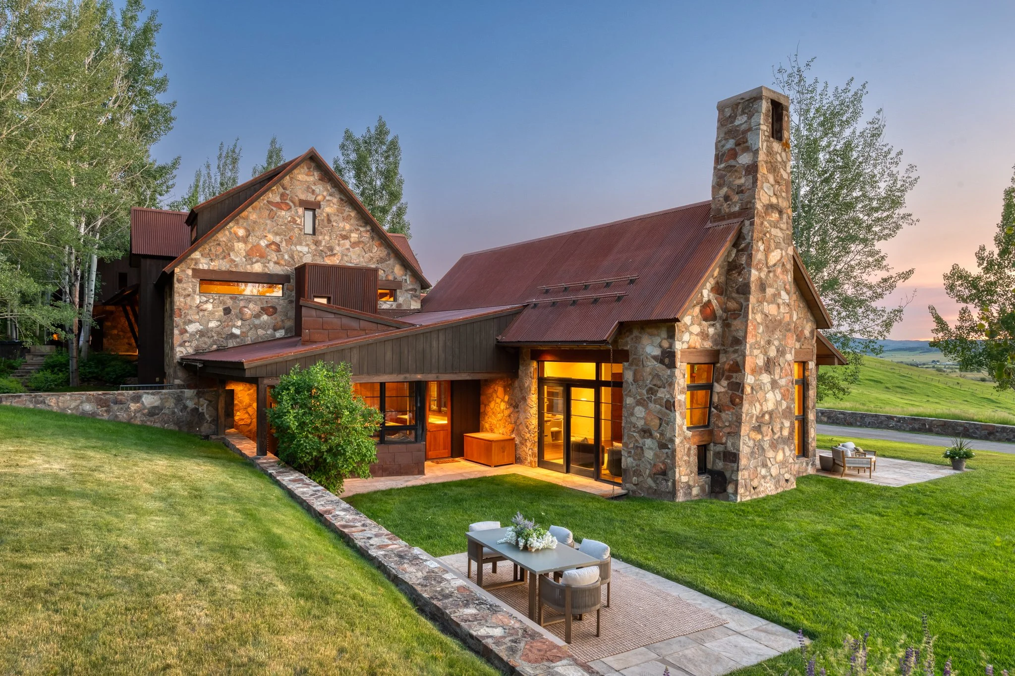 A stone and wood house with a red metal roof, illuminated warmly inside, surrounded by a green lawn and tall trees, with an outdoor sitting area and a scenic countryside in the background at sunset.