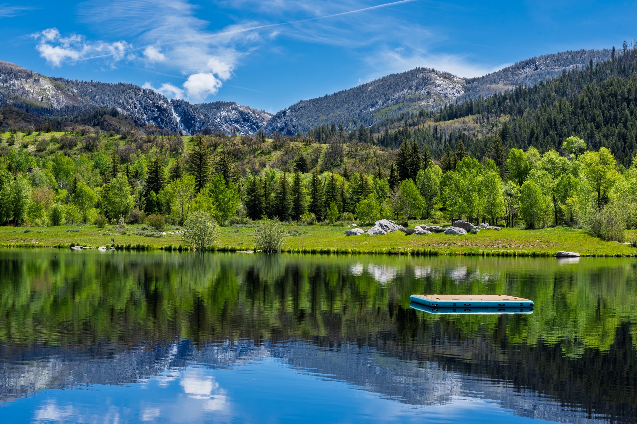 A peaceful mountain landscape with snow-capped peaks, a lush green forest, and a calm lake reflecting the sky and trees. A blue and white paddleboard is floating on the lake in the foreground.