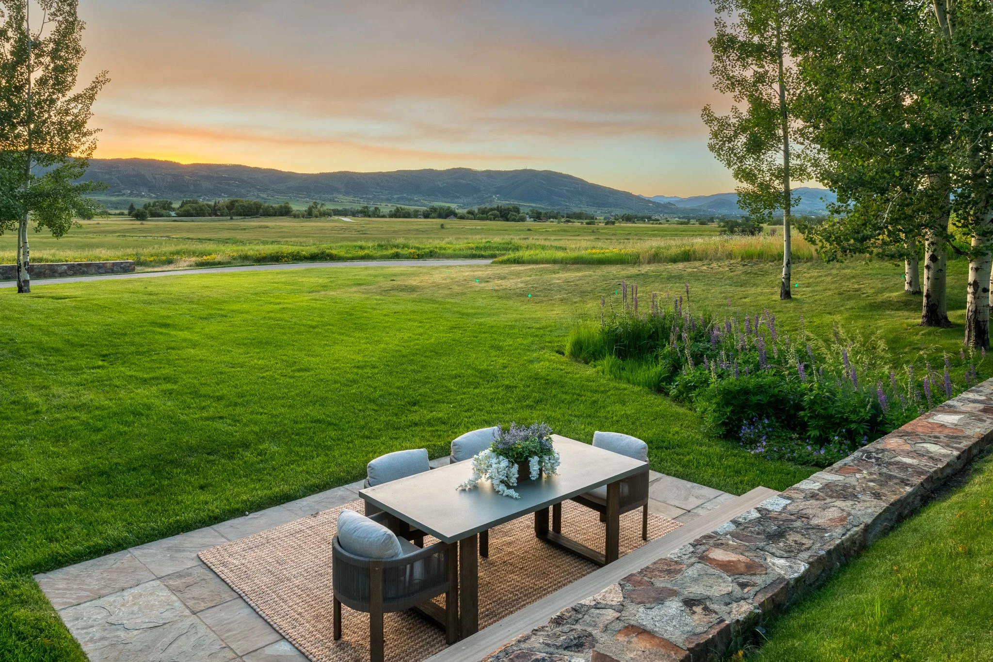 A backyard patio with a table and six chairs, overlooking a lush green field and distant mountains at sunset.