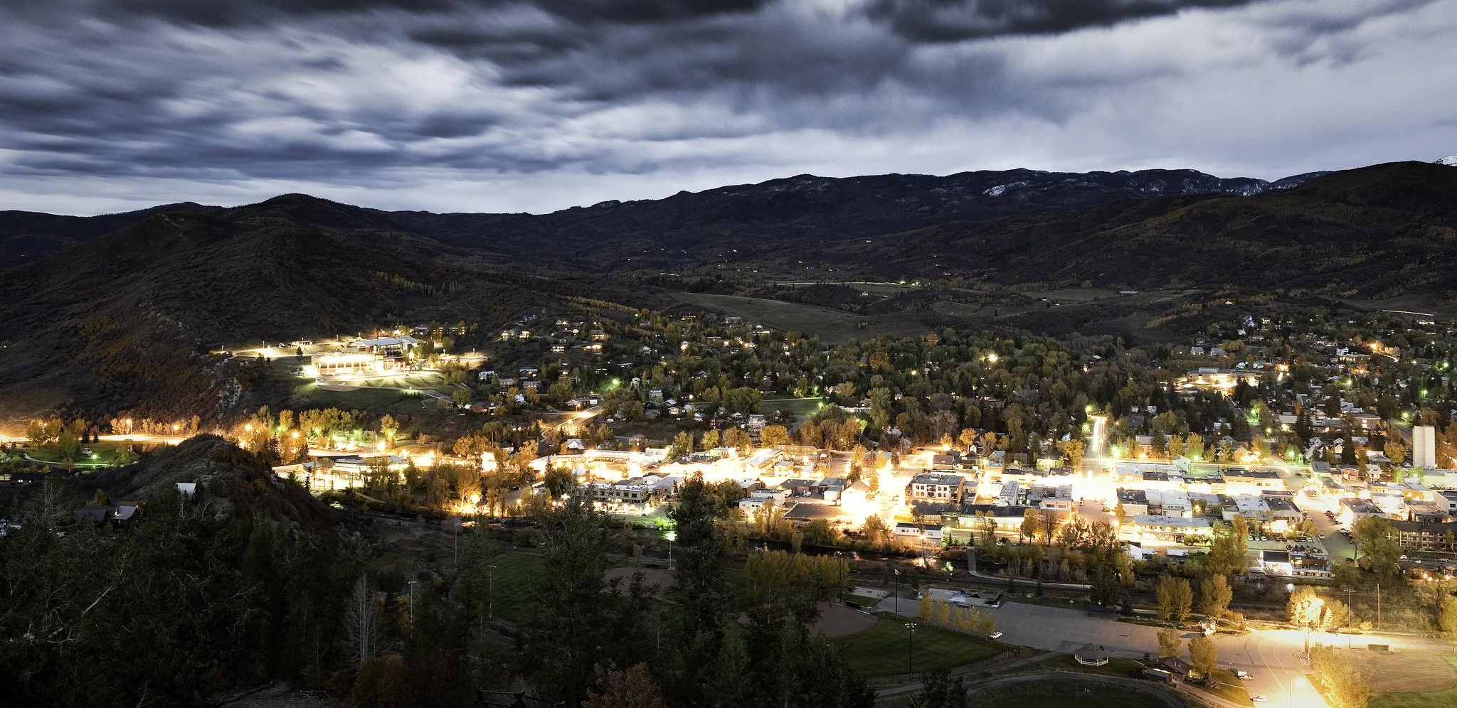 A town at night with brightly lit buildings and streets, surrounded by hills and mountains under a cloudy sky.