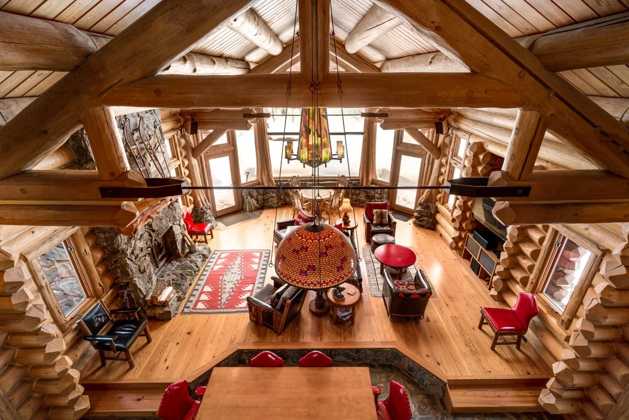 Interior view of a log cabin living room with wooden beams, furniture including chairs, sofas, and tables, a stone fireplace with logs, and large windows letting in natural light.