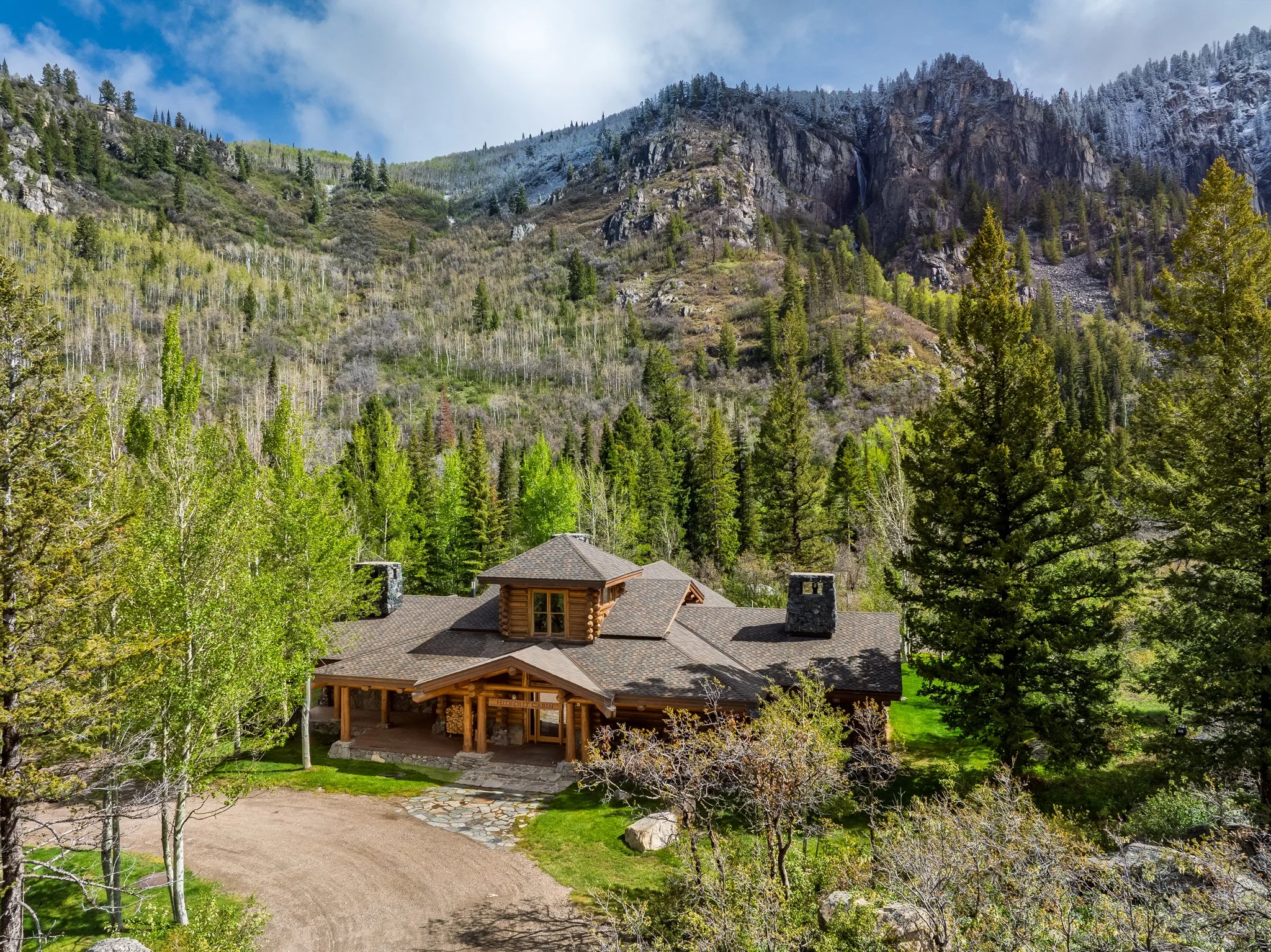A log cabin with a tiled roof surrounded by trees and a gravel driveway, set against a mountain landscape with green and snow-capped peaks.