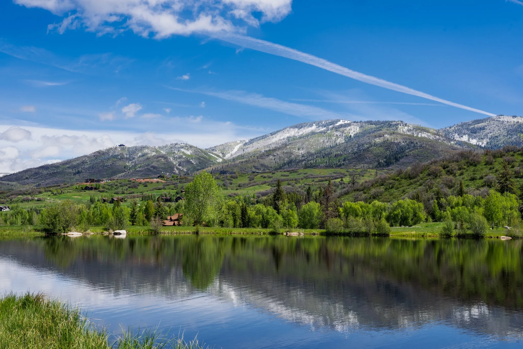 Mountain landscape with green trees, a lake in the foreground, and snowy peaks under a blue sky with clouds and contrails.