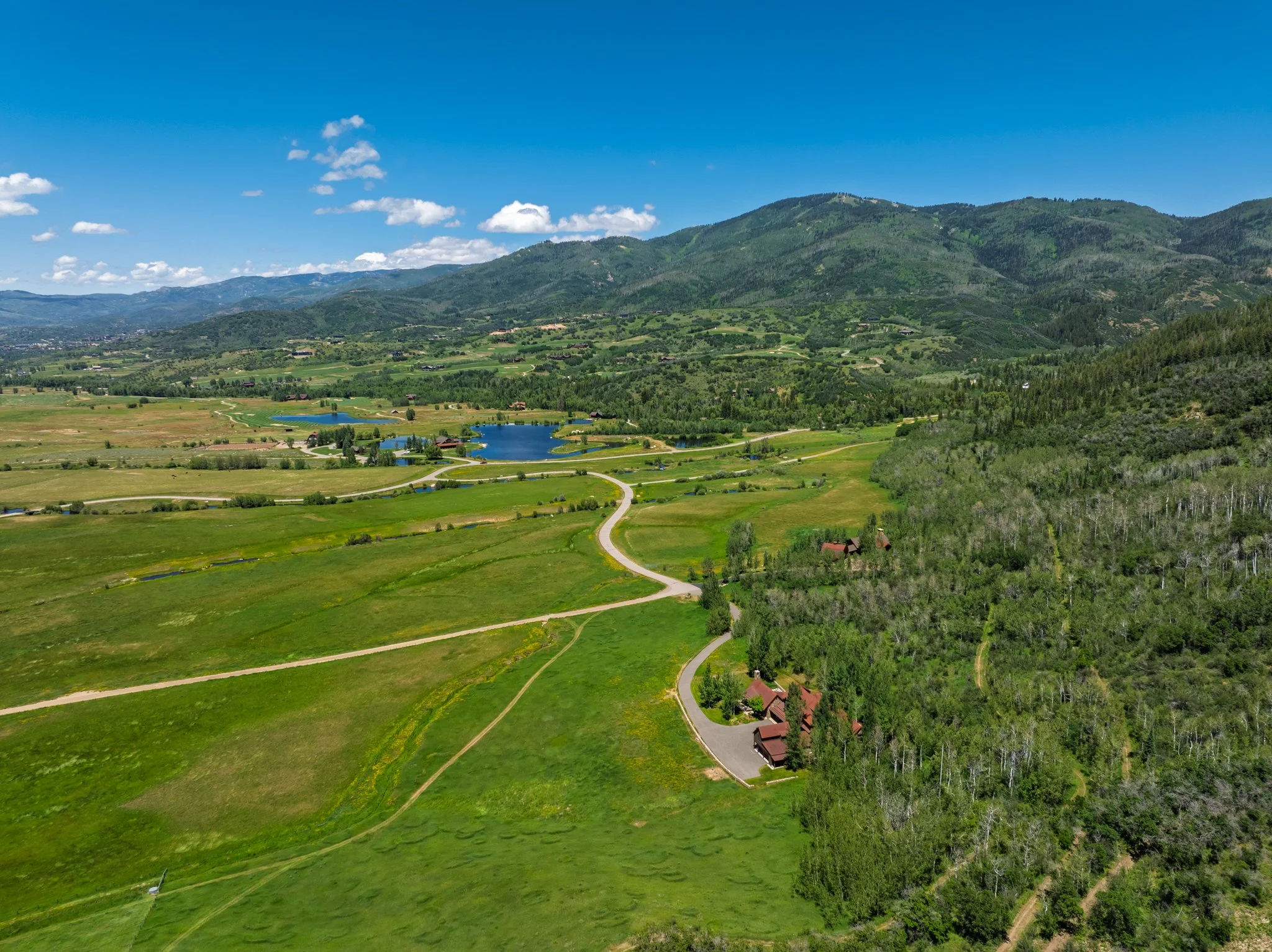 Scenic view of green rolling hills, small lakes, and mountains under a blue sky with scattered clouds.