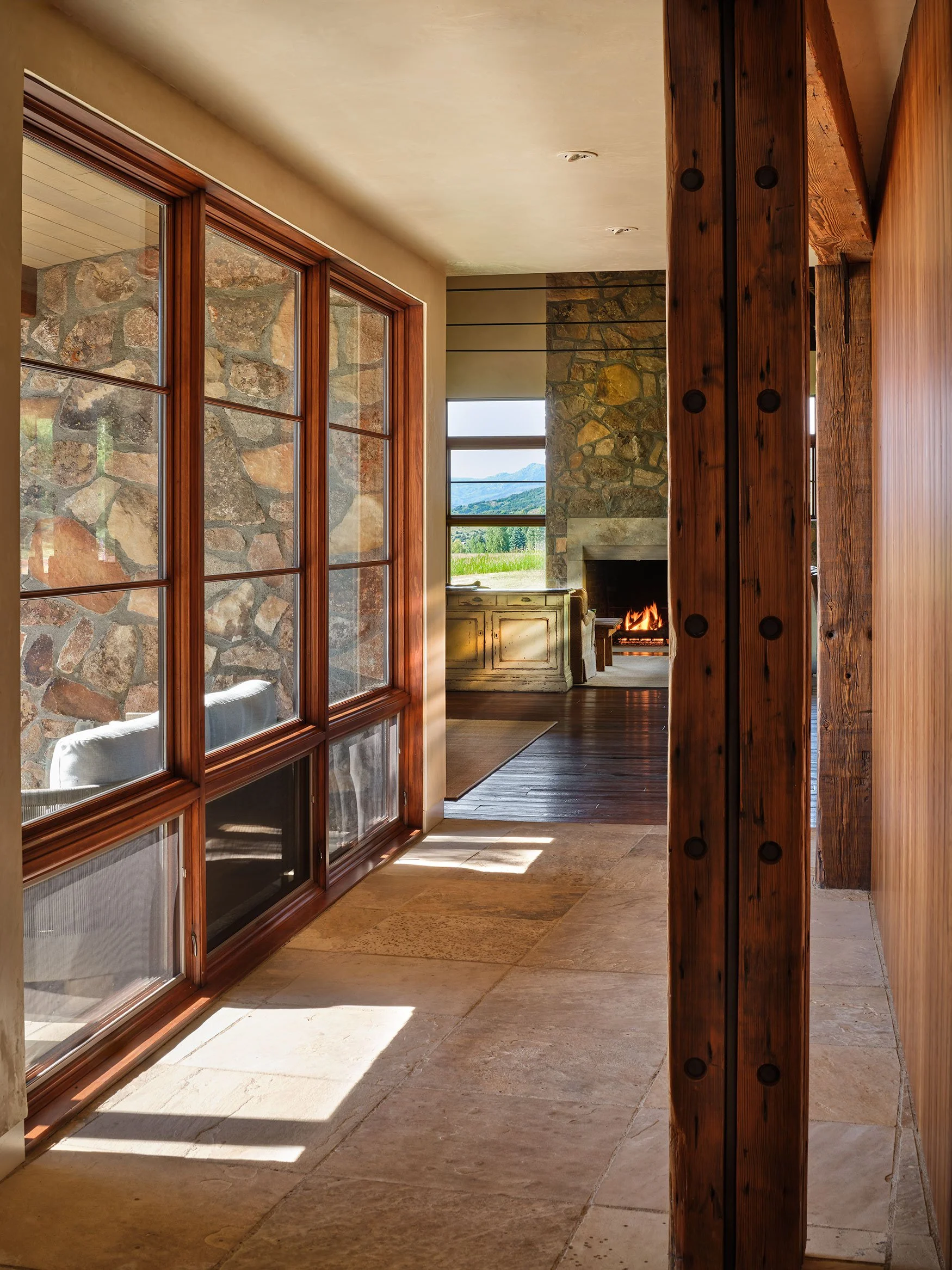 A hallway featuring large wooden-framed glass windows, a stone wall, and a rustic wooden beam with metal bolts. In the background, a cozy fireplace and a window showing a mountain landscape are visible.