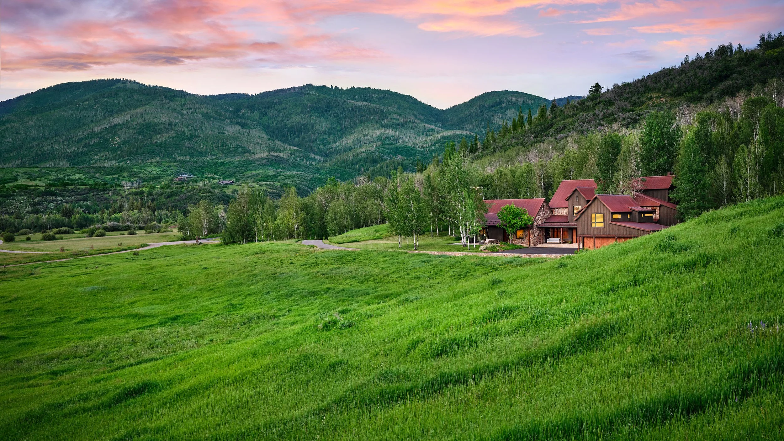 Scenic view of a rural house surrounded by green grass and trees, with forested mountains in the background under a pink and blue cloudy sky.