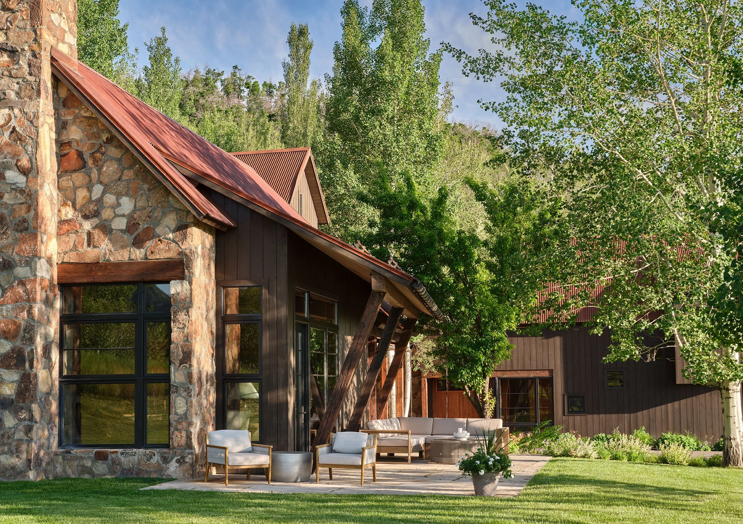 Exterior view of a house with a stone and wood facade, large windows, a copper roof, and an outdoor seating area with chairs and a table, surrounded by green trees and lawn under a partly cloudy sky.