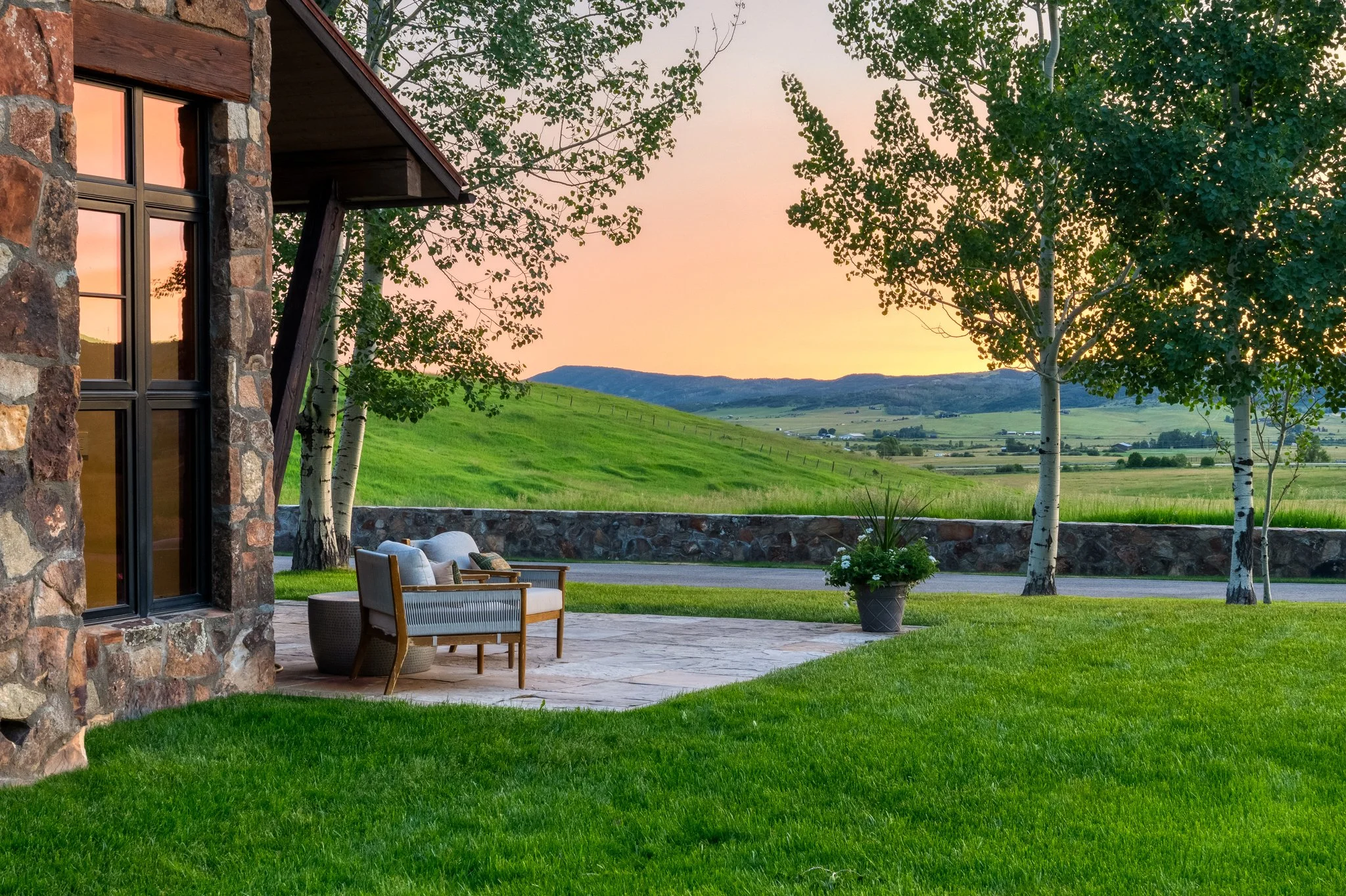 A stone house with large windows and a patio, overlooking a lush green landscape with trees and rolling hills at sunset.