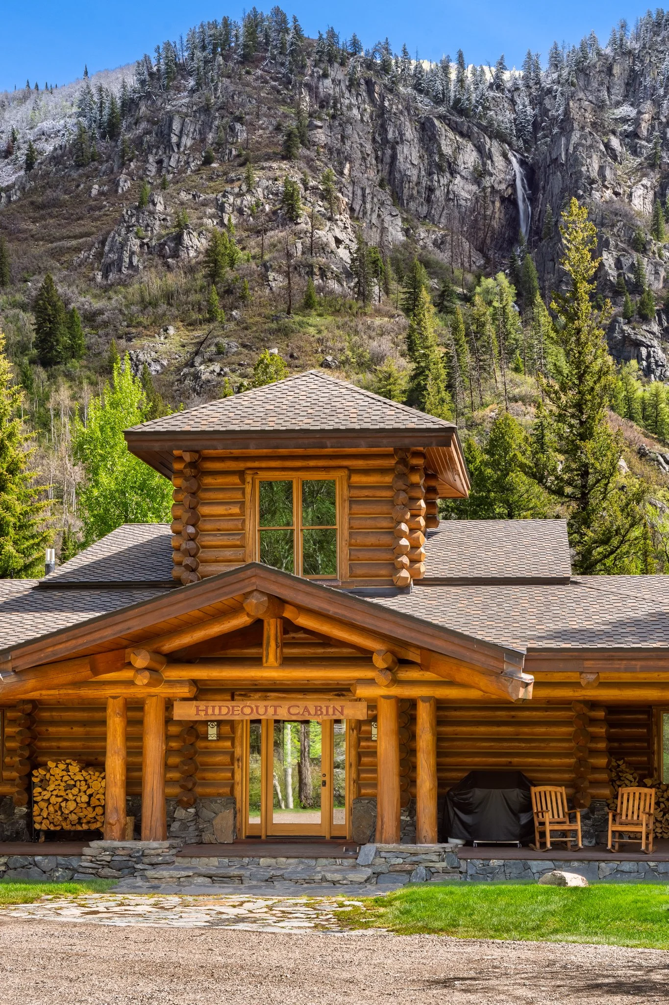 A wooden cabin with a sign that reads 'HIDEOUT CABIN' at the entrance, surrounded by trees and set against a mountainous backdrop with a waterfall and snowy peaks.