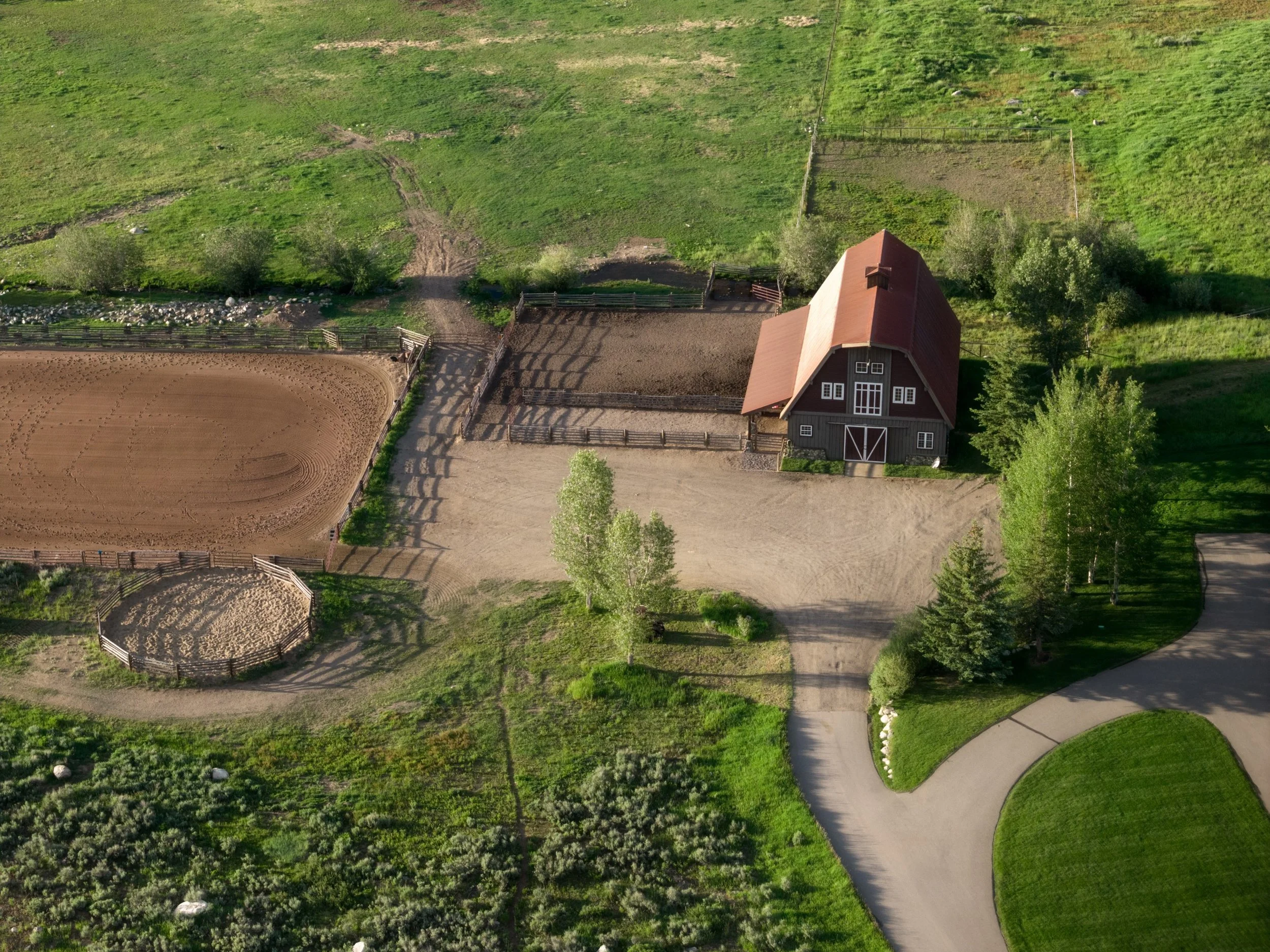 Aerial view of a farm with a barn and fenced areas, surrounded by green grass, trees, and a dirt driveway.