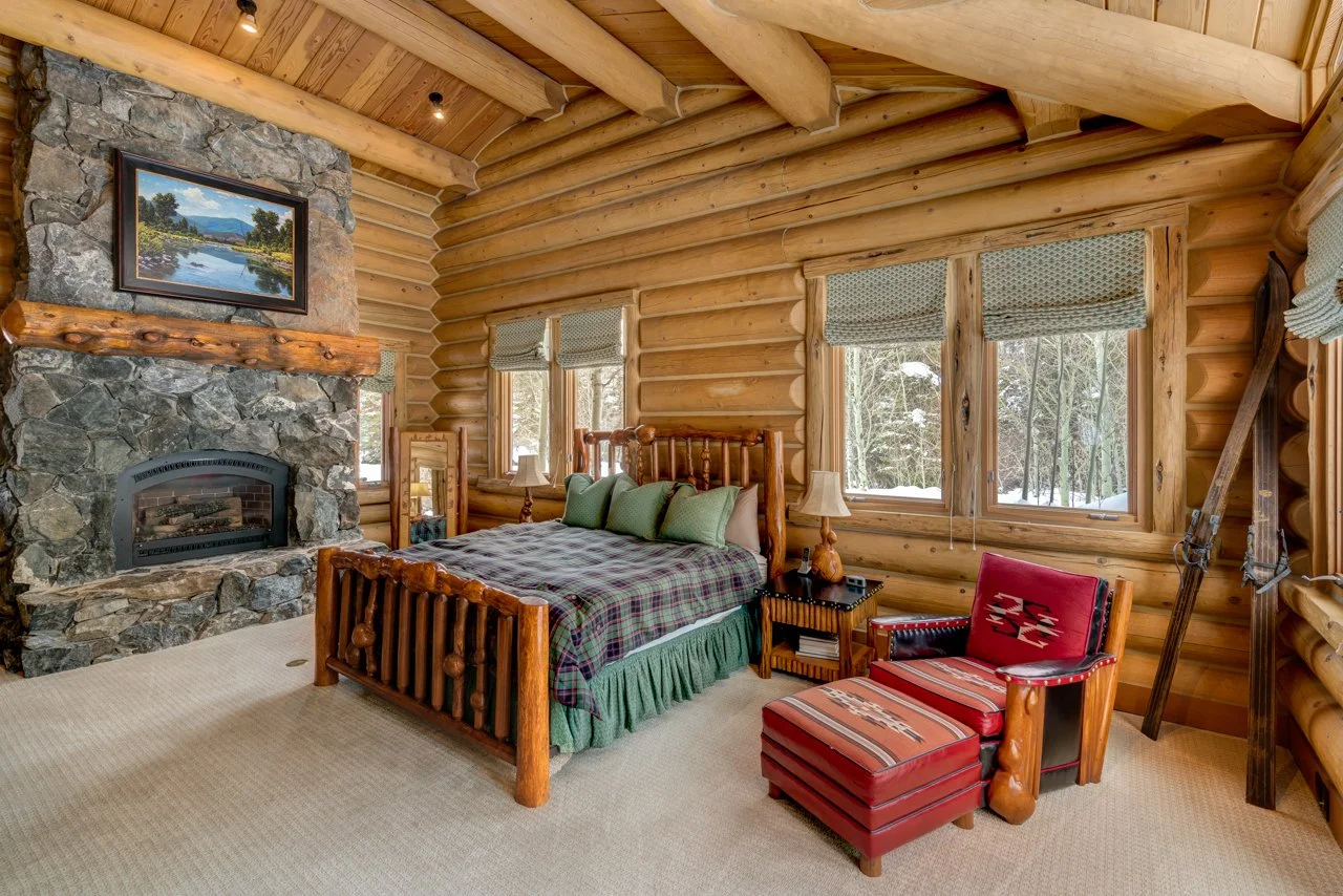 A cozy log cabin bedroom with a stone fireplace, wooden walls and ceiling, a bed with green pillows, side table with lamps, a red armchair with an ottoman, and snow outside the windows.