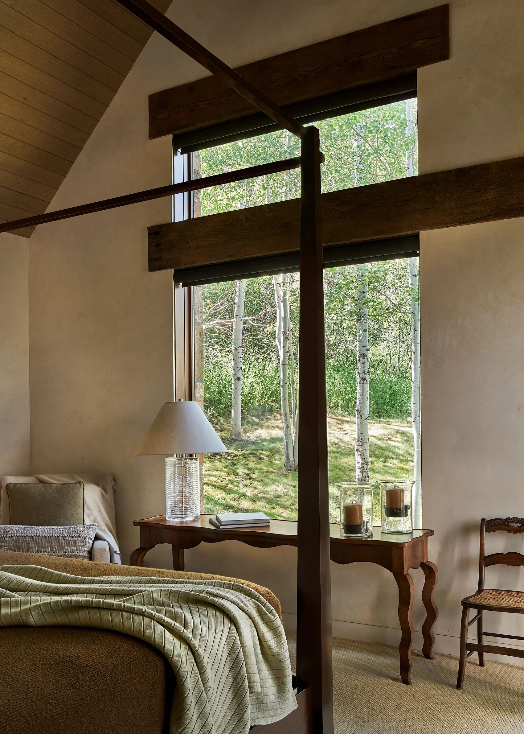 A cozy bedroom scene with a bed, nightstand, lamp, and a window looking out to a wooded area with trees.