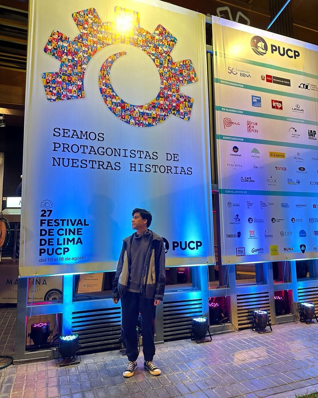 Una persona joven de pie frente a carteles en un festival de cine en Lima, Perú, promocionando la edición número 27 del Festival de Cine de Lima PUCP, con fechas del 10 al 18 de agosto.