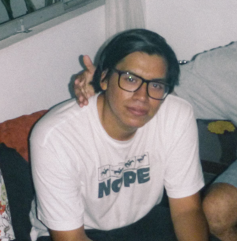 Joven con gafas, cabello oscuro, sentado en una cama, camiseta blanca con texto y gráficos, en un ambiente interior.