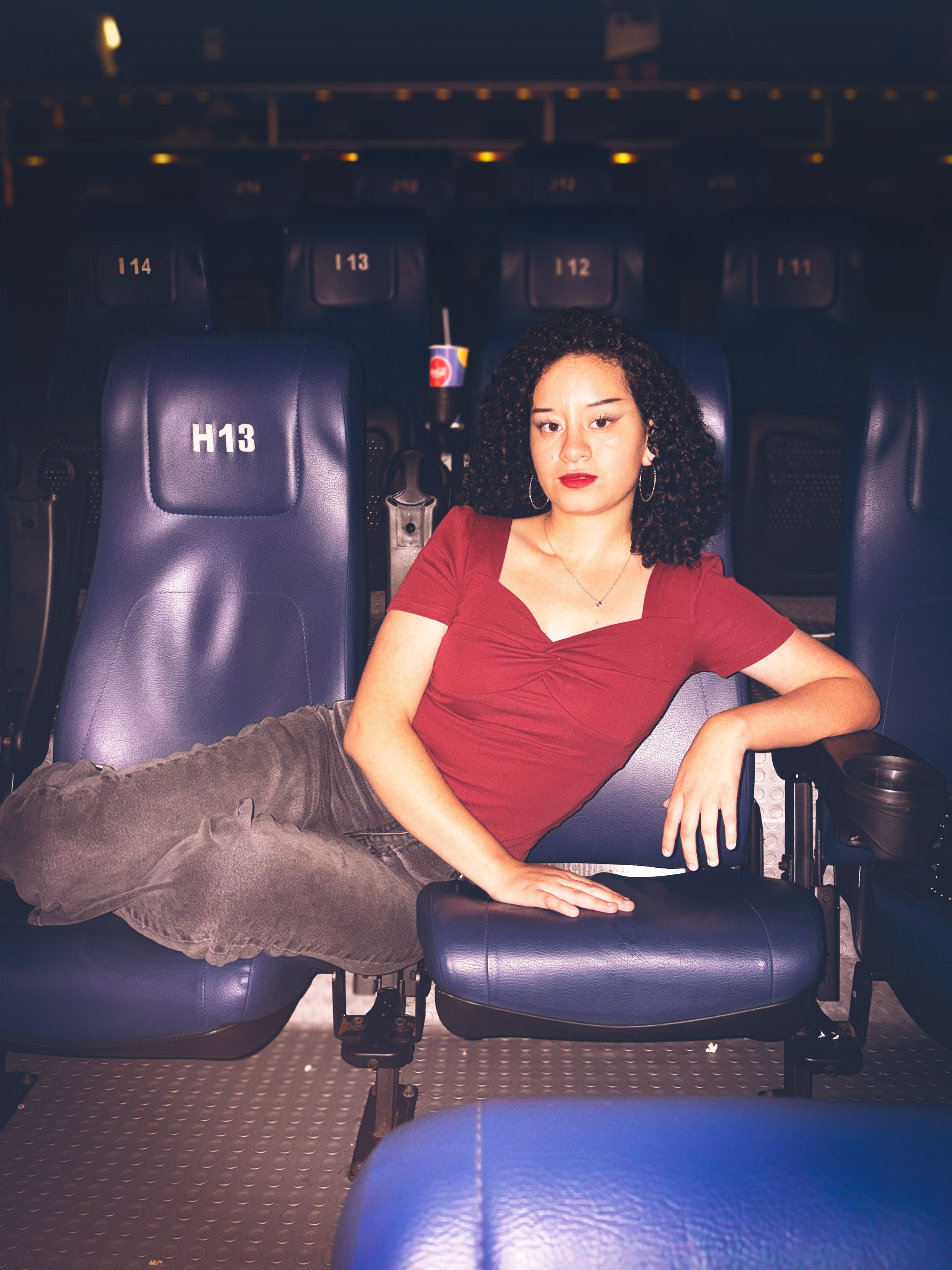 Mujer con cabello rizado, vestida con blusa roja, recostada en una fila de asientos en un cine o teatro oscuro, con una bebida en un vaso con popote en un asiento detrás.