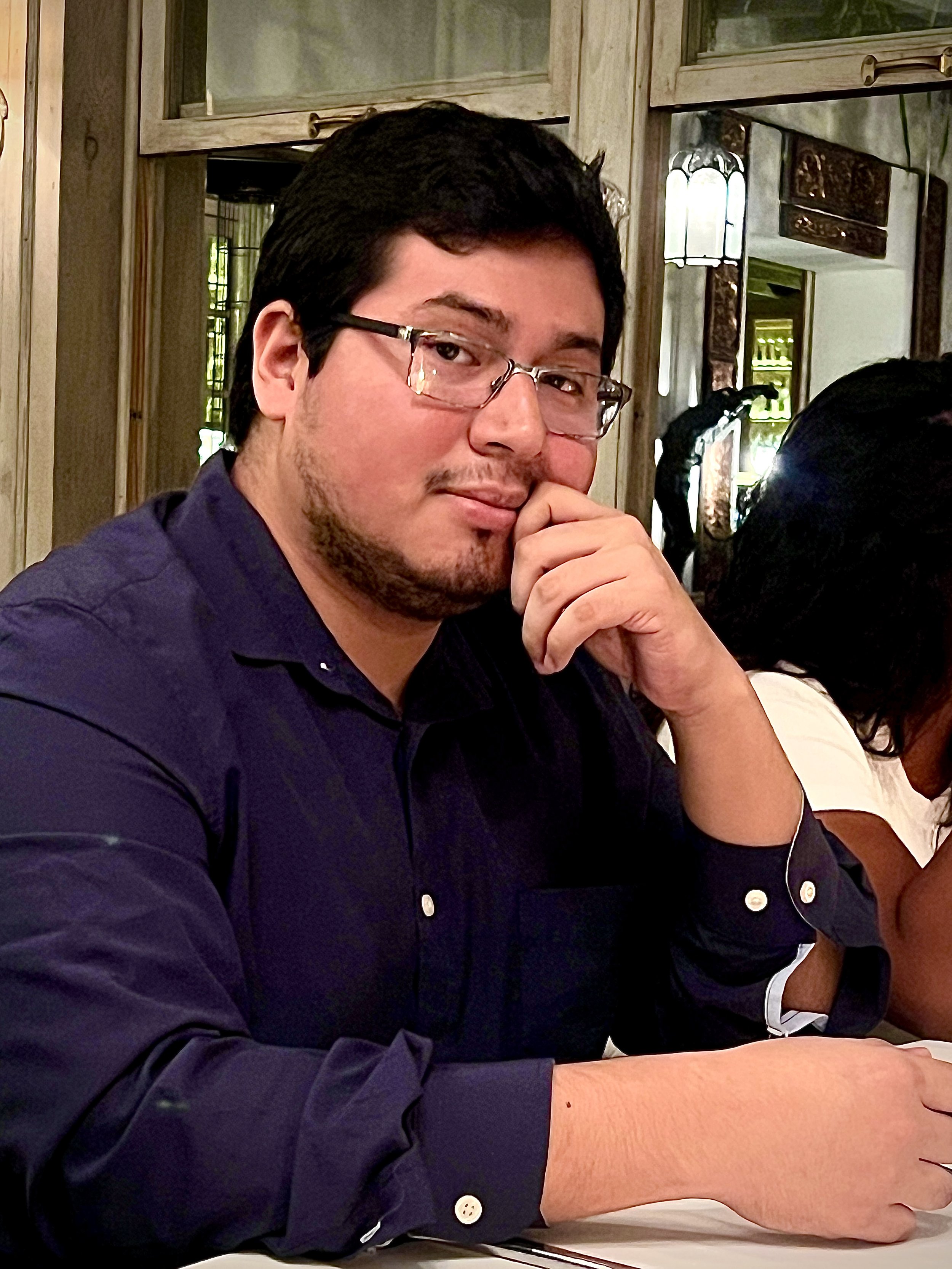 Un hombre con gafas de cabello oscuro y piel clara, sentado en una mesa, mira hacia la cámara con una expresión pensativa y la mano en su rostro.