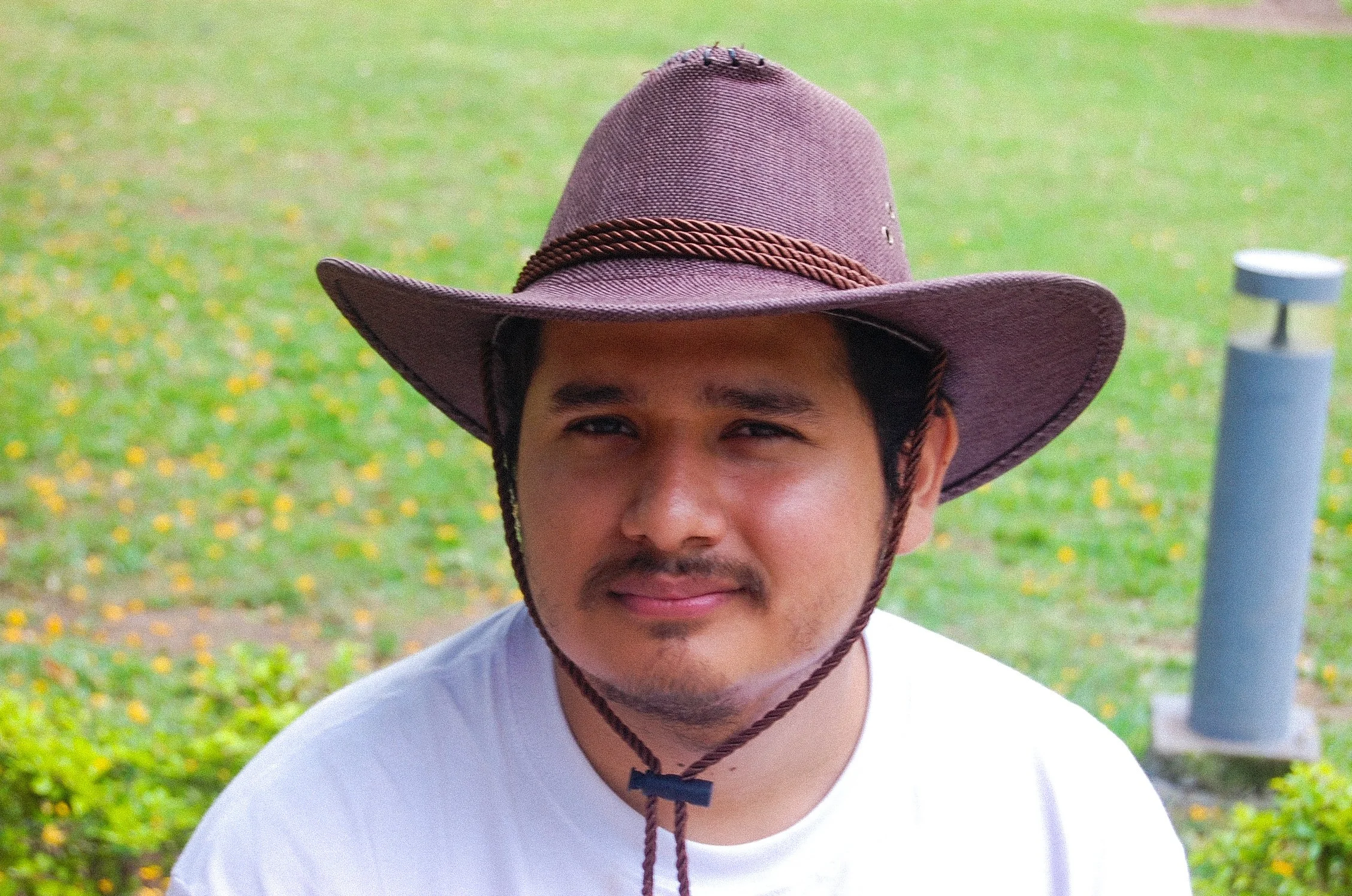 Joven sonriendo con sombrero de vaquero en un parque con césped y flores amarillas.