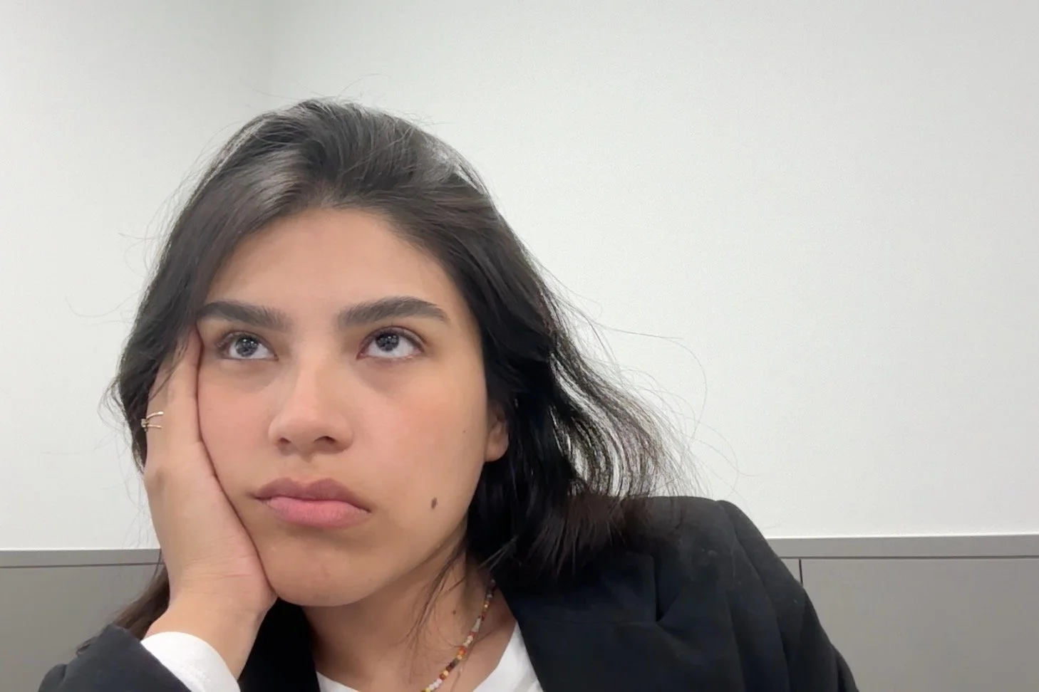 Retrato de una joven mujer con expresión pensativa, sosteniendo su rostro con una mano, lleva una chaqueta negra y un collar, con fondo de pared blanca.