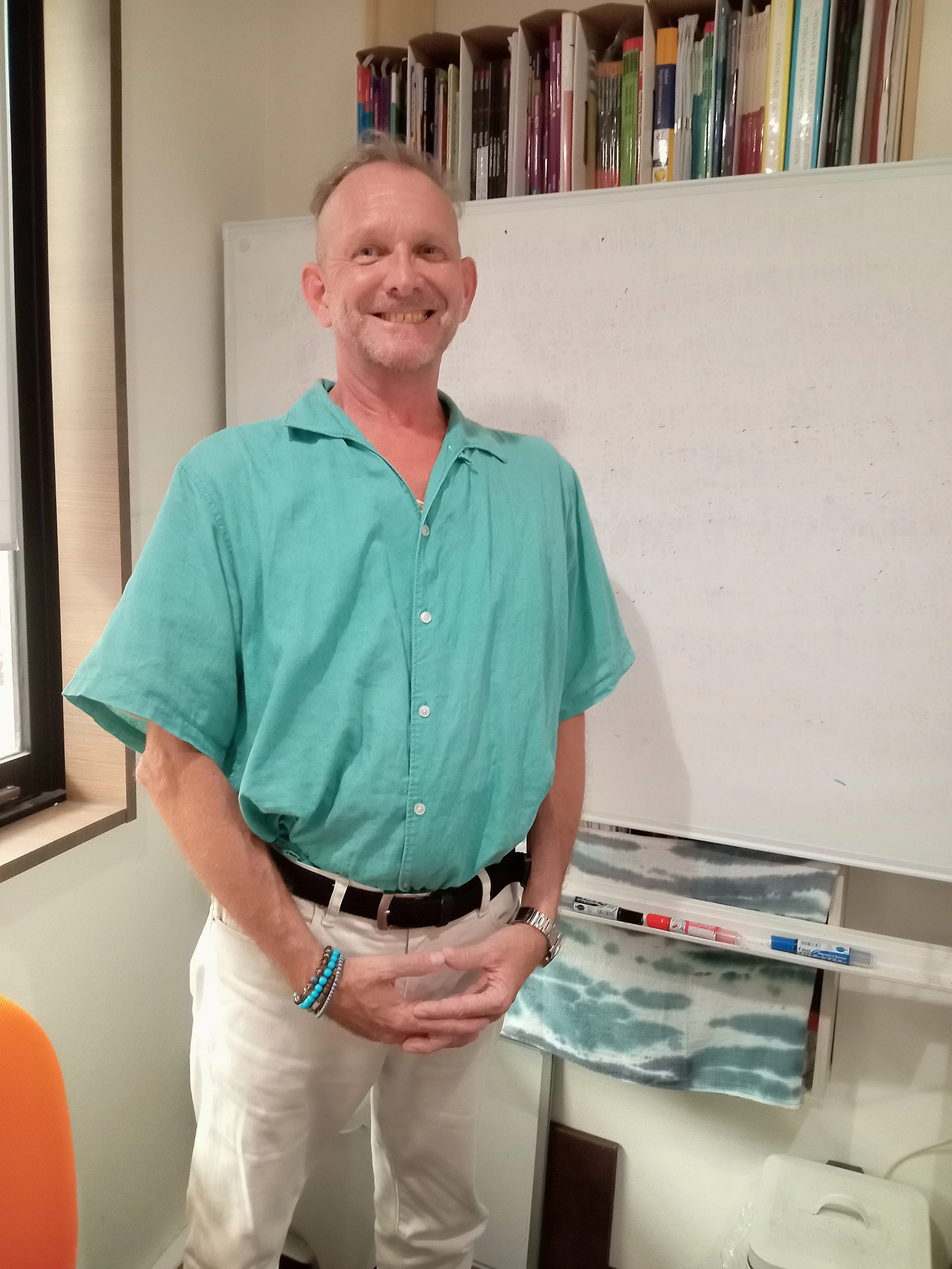 A smiling man wearing a turquoise short-sleeve collared shirt and white pants, standing in front of a whiteboard with books on shelves behind him, holding his hand in a gesture.
