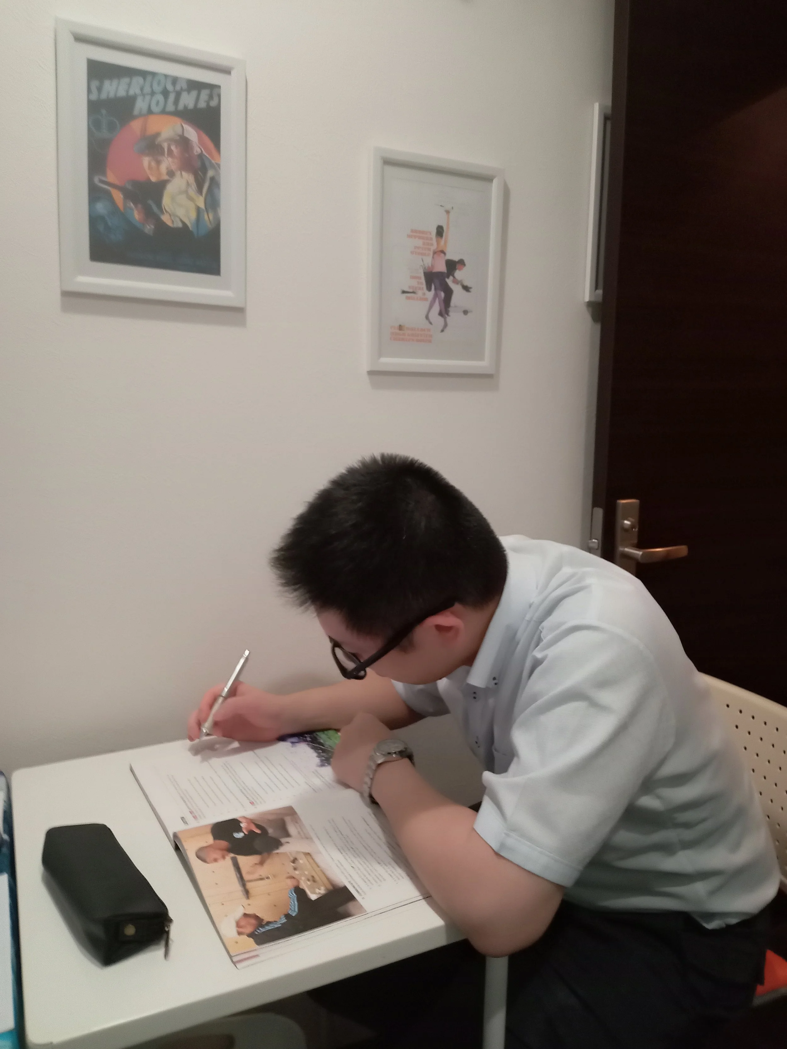 A person with short dark hair and glasses, wearing a white shirt, sitting at a white table, writing on paper with a pen, on a white chair in a room with white walls. There are two framed posters on the wall, one of Sherlock Holmes and the other with animated characters. On the table are a magazine, a black pen case, and a black speaker.