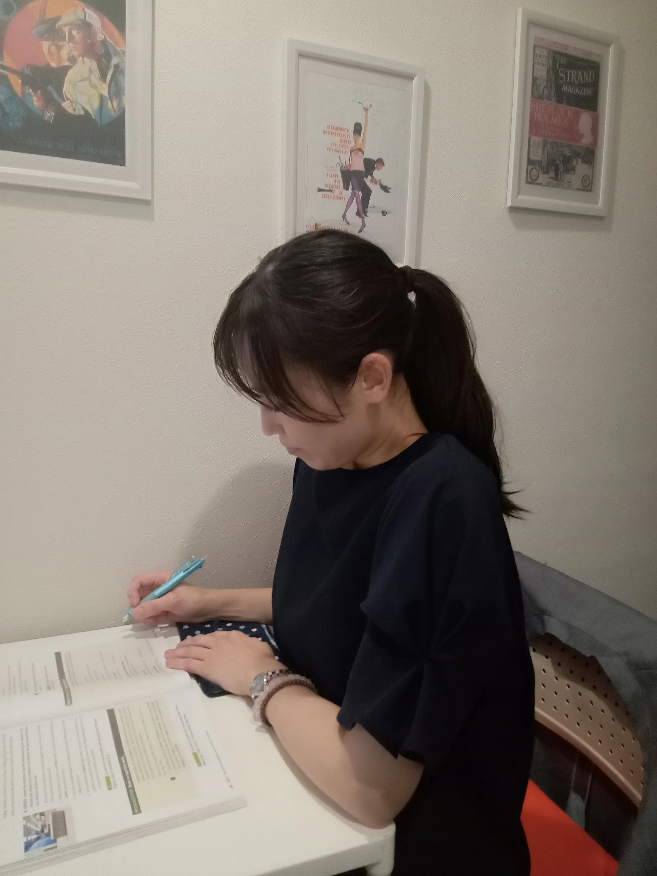 A woman with dark hair tied in a ponytail, wearing a black T-shirt and a beaded bracelet, is sitting at a white table reading a large brochure or catalog, and holding a pen. On the wall behind her, there are three framed posters or prints.
