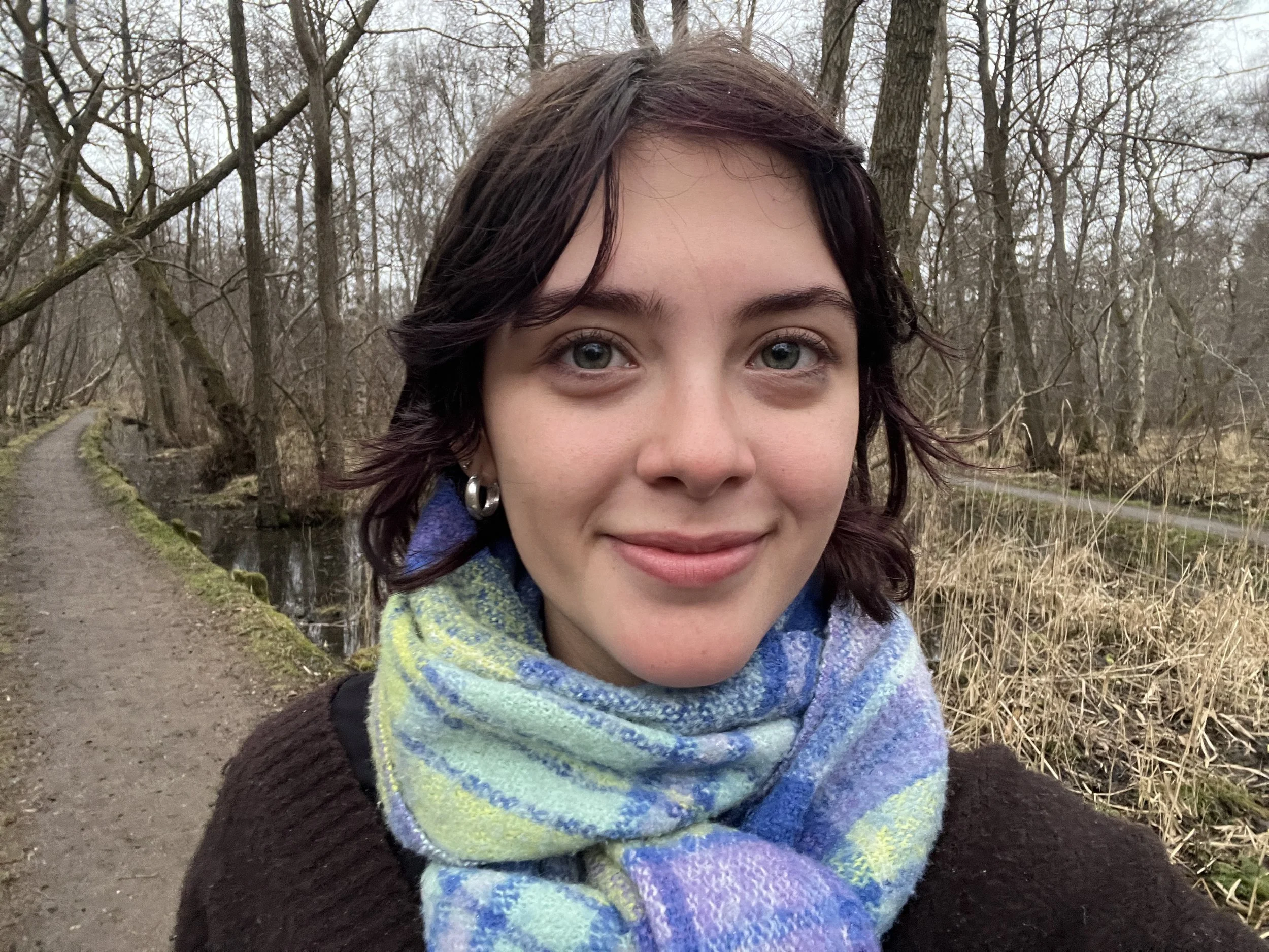 Close-up of a woman with curly dark hair, wearing a multicolored scarf, standing outdoors in a wooded area during overcast weather.