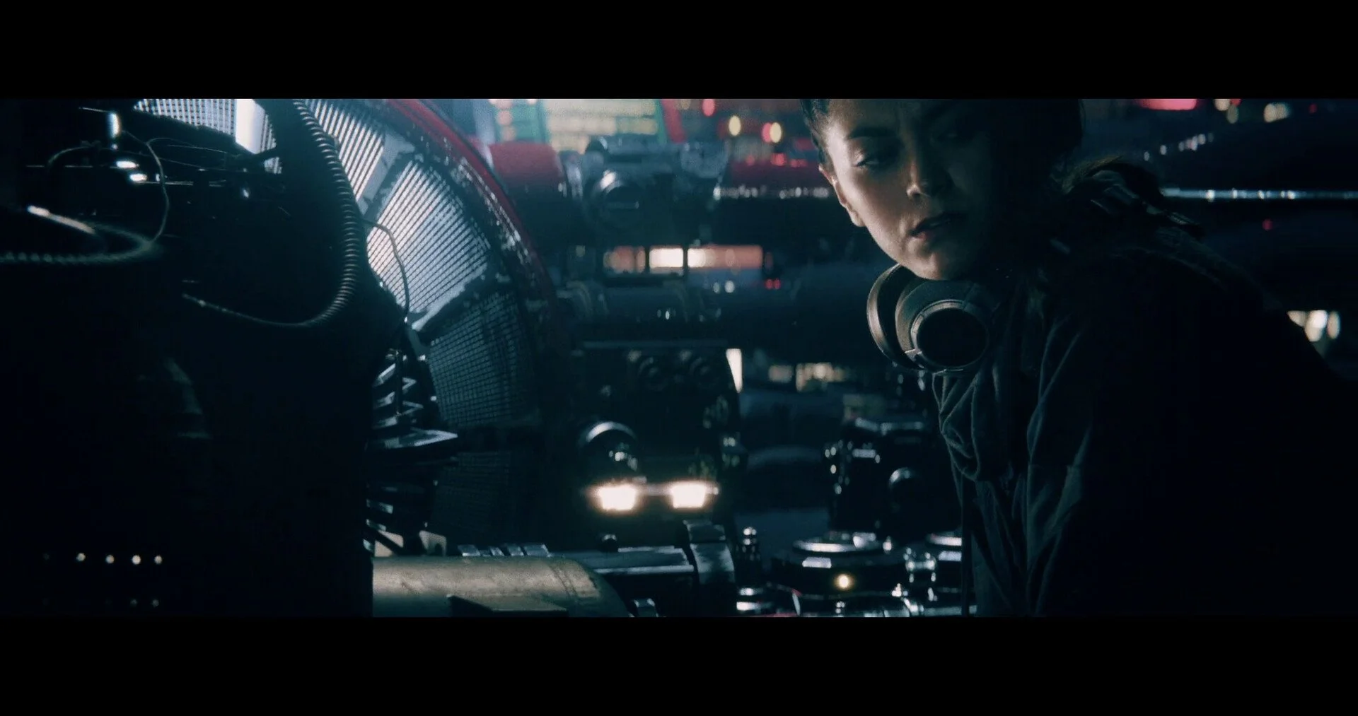 A woman with headphones around her neck leans inside a vehicle, possibly a bus or a van, with city lights and traffic behind her at night.