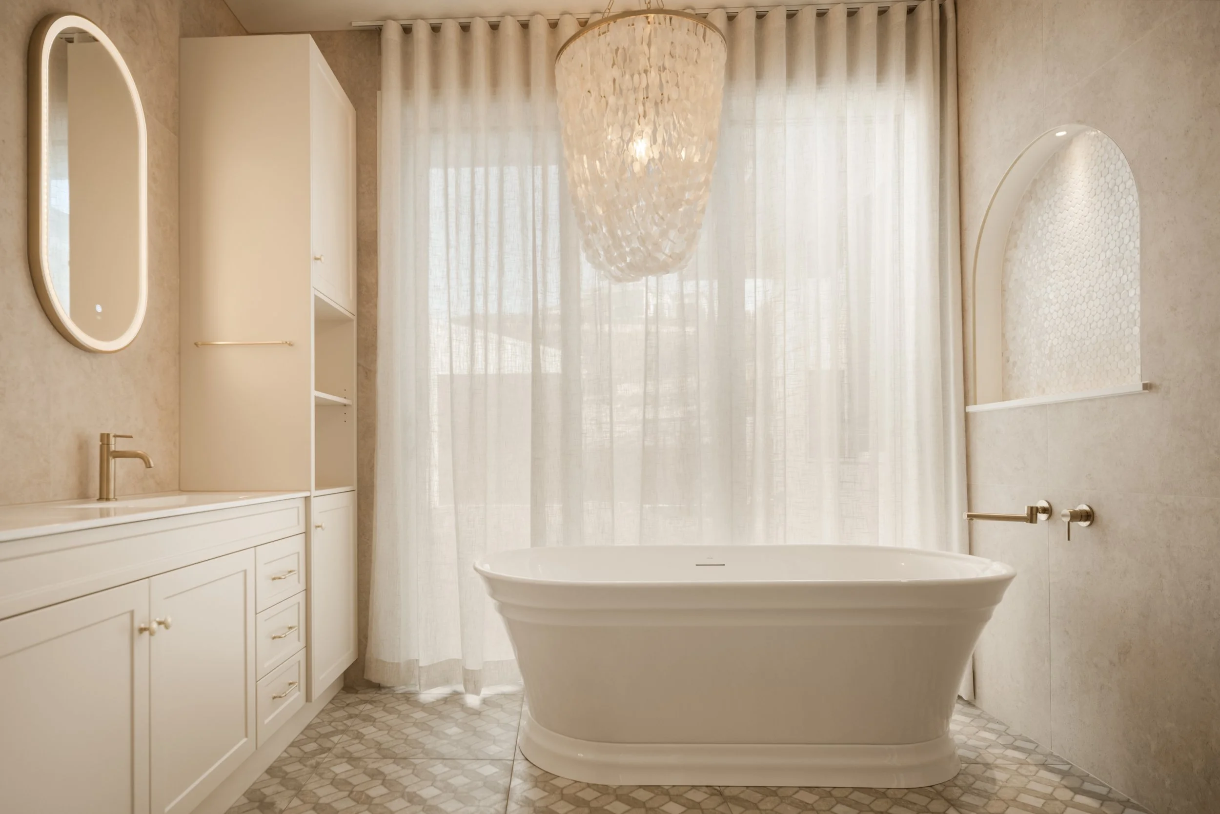 Bright bathroom with a freestanding tub, white cabinets, a large window with sheer curtains, an oval mirror, and a modern chandelier.