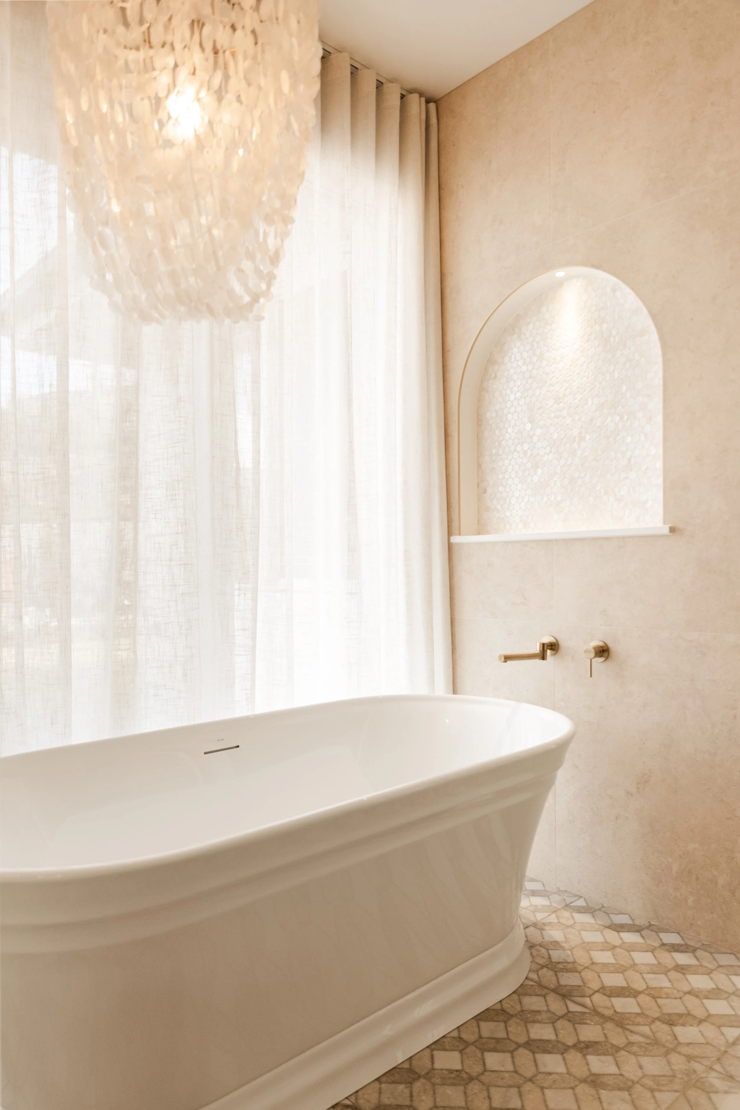 A bathroom with a freestanding white bathtub, beige stone walls, a textured alcove with tile, a window with sheer curtains, and a chandelier with shell-like design.