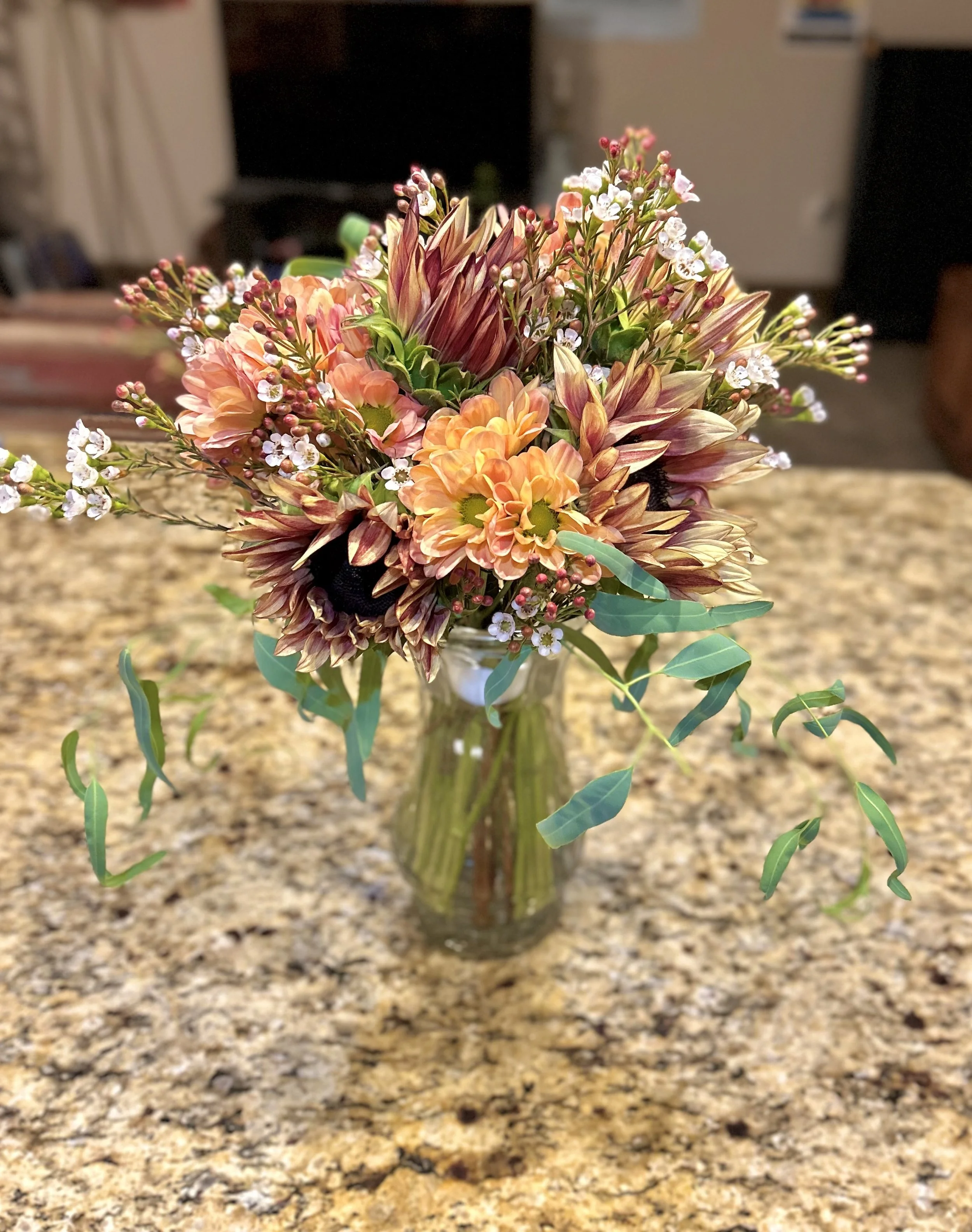 A colorful bouquet of flowers in a glass vase on a granite countertop. The bouquet includes various flowers such as orange and yellow chrysanthemums and sunflowers, eucalyptus leaves, and small white waxflowers.