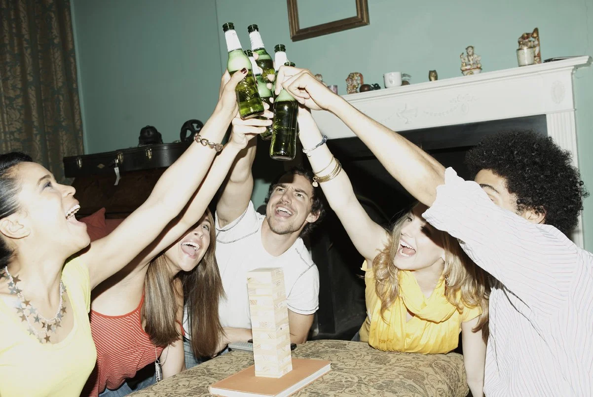 A group of six friends celebrating with drinks raised together in a toast indoors, sitting around a table with a Jenga game on it.
