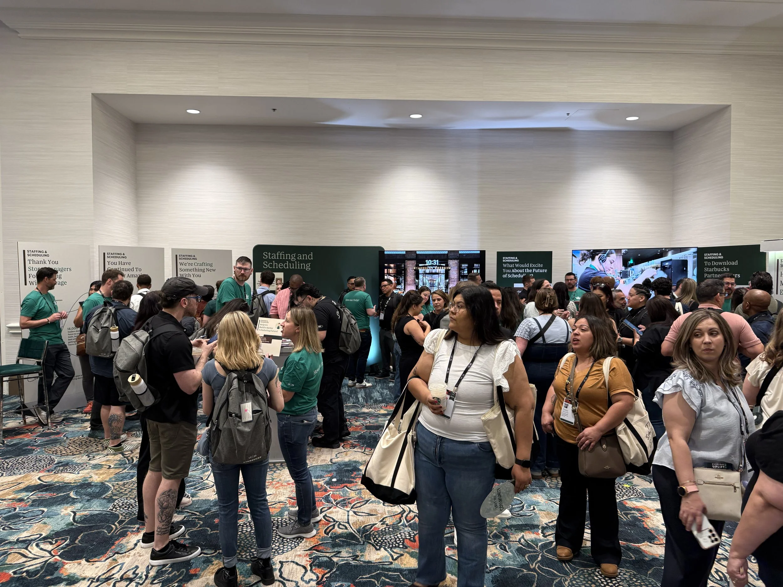 Crowd of people at an indoor event, standing near tables and booths with posters and signs about staffing, scheduling, and new developments.