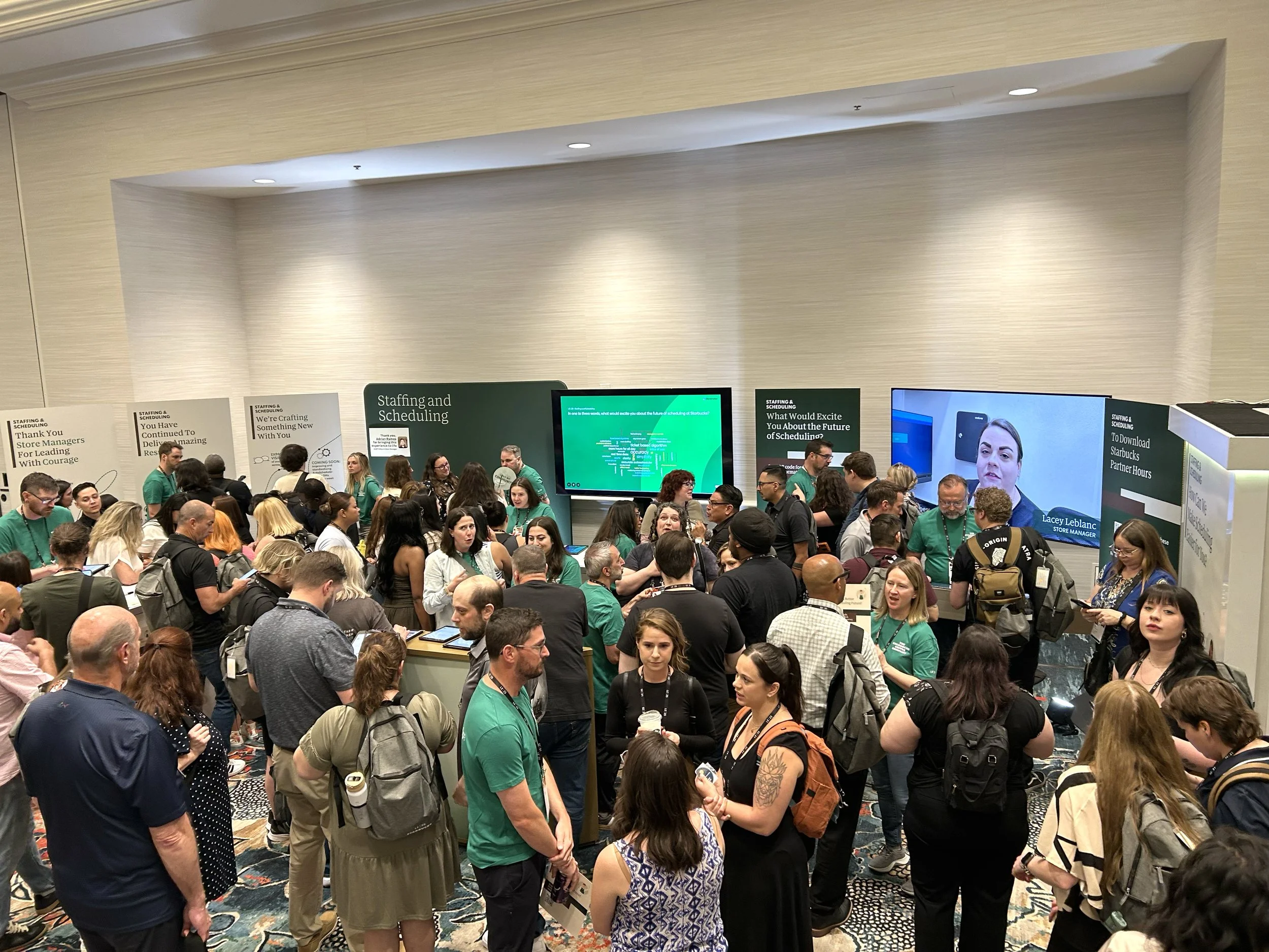 A crowded conference registration area with many people waiting in line and gathered around. There are several posters and screens about staffing, scheduling, and store management on the walls.