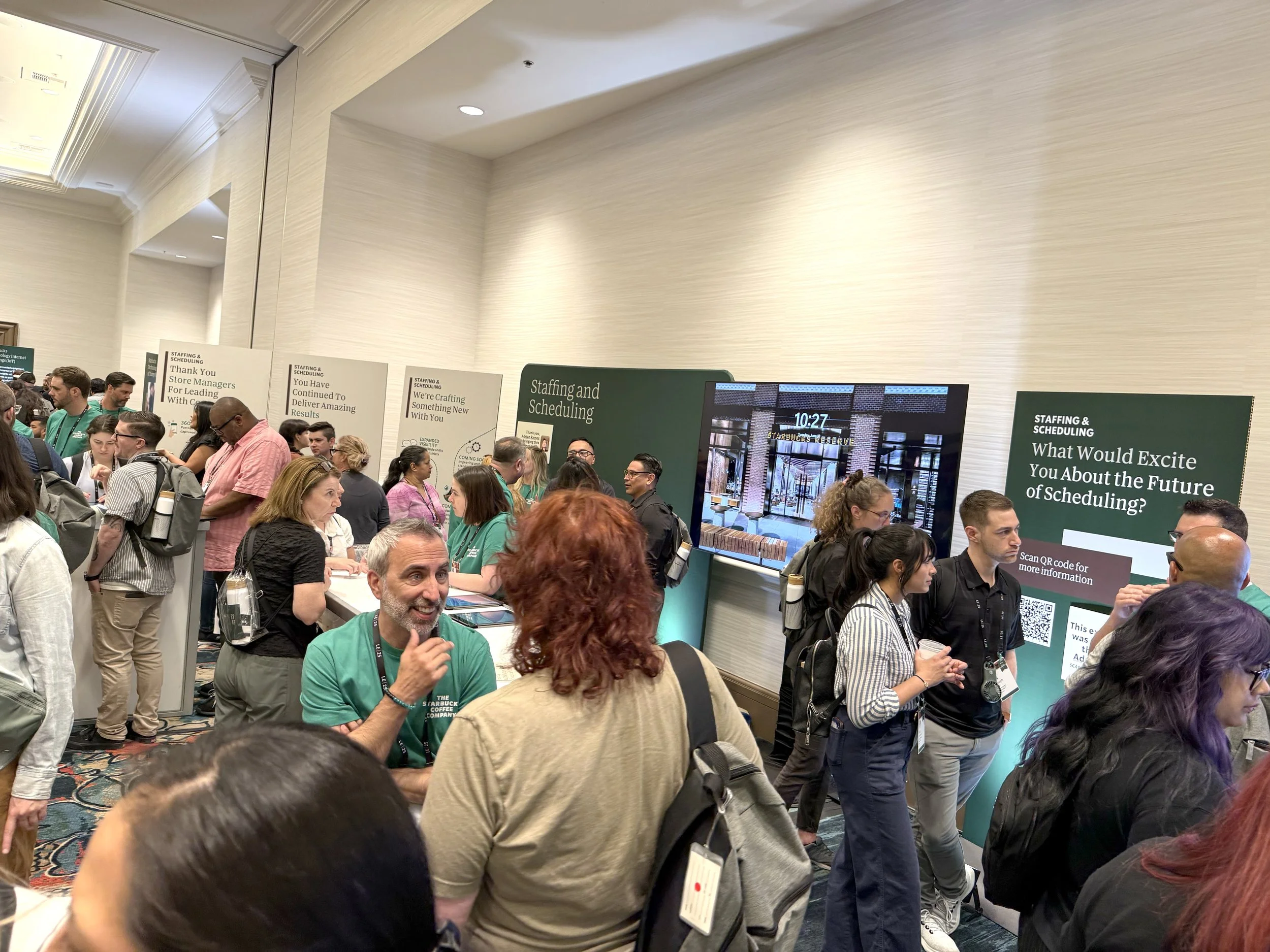 A crowded conference at the Starbucks Leadership conference with attendees near booths and large screens displaying information about staffing and scheduling.