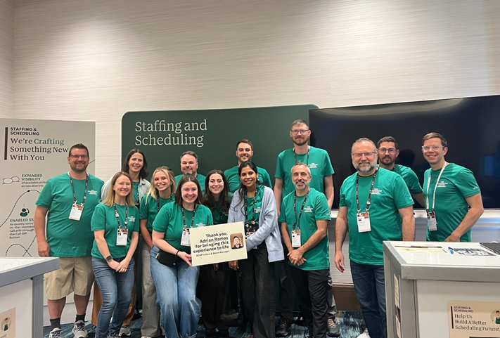 Group of 14 people standing in front of a green wall that says 'Staffing and Scheduling' at a conference or event, some wearing name tags, posing for a group photo.