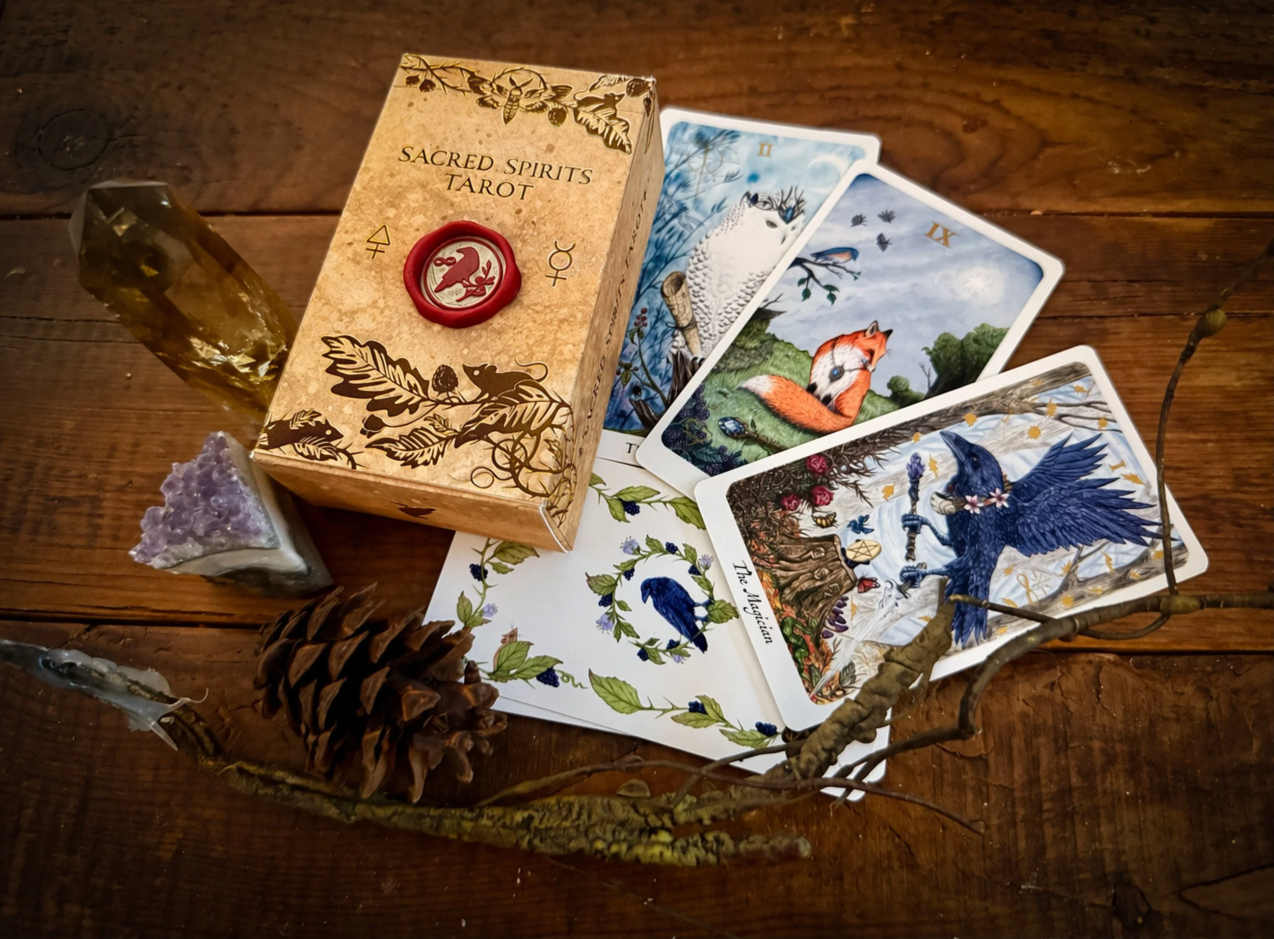 A tarot card deck called Sacred Spirits Tarot, with three cards fanned out on a wooden surface. The images on the cards include a white owl, a fox, and a dark winged figure with a staff. There is also a pyramid-shaped crystal, a pine cone, and a dried branch on the surface.