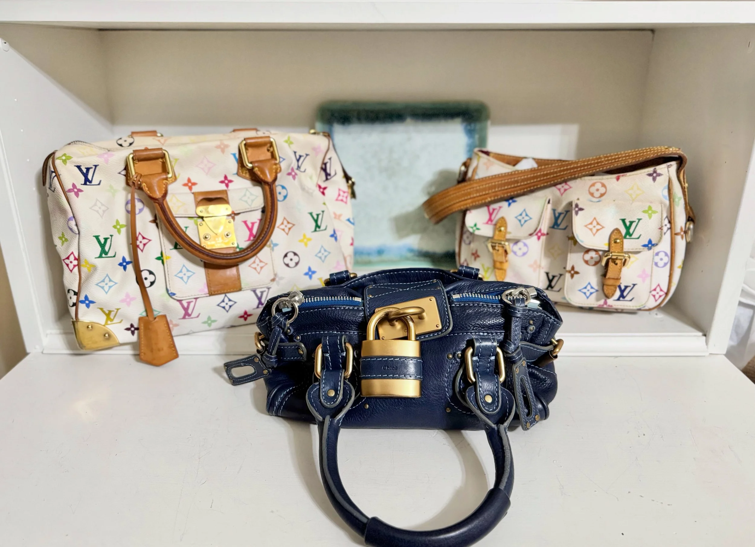 Display of designer handbags on a white shelf, including colorful Louis Vuitton bags with multicolored monogram pattern, and a dark blue leather handbag in front.