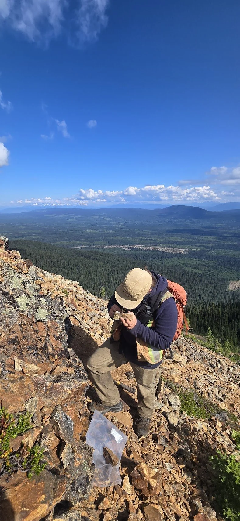 SFD Consulting's Jared Liimu examines a sample while standing on the side of a steep, rocky slope.