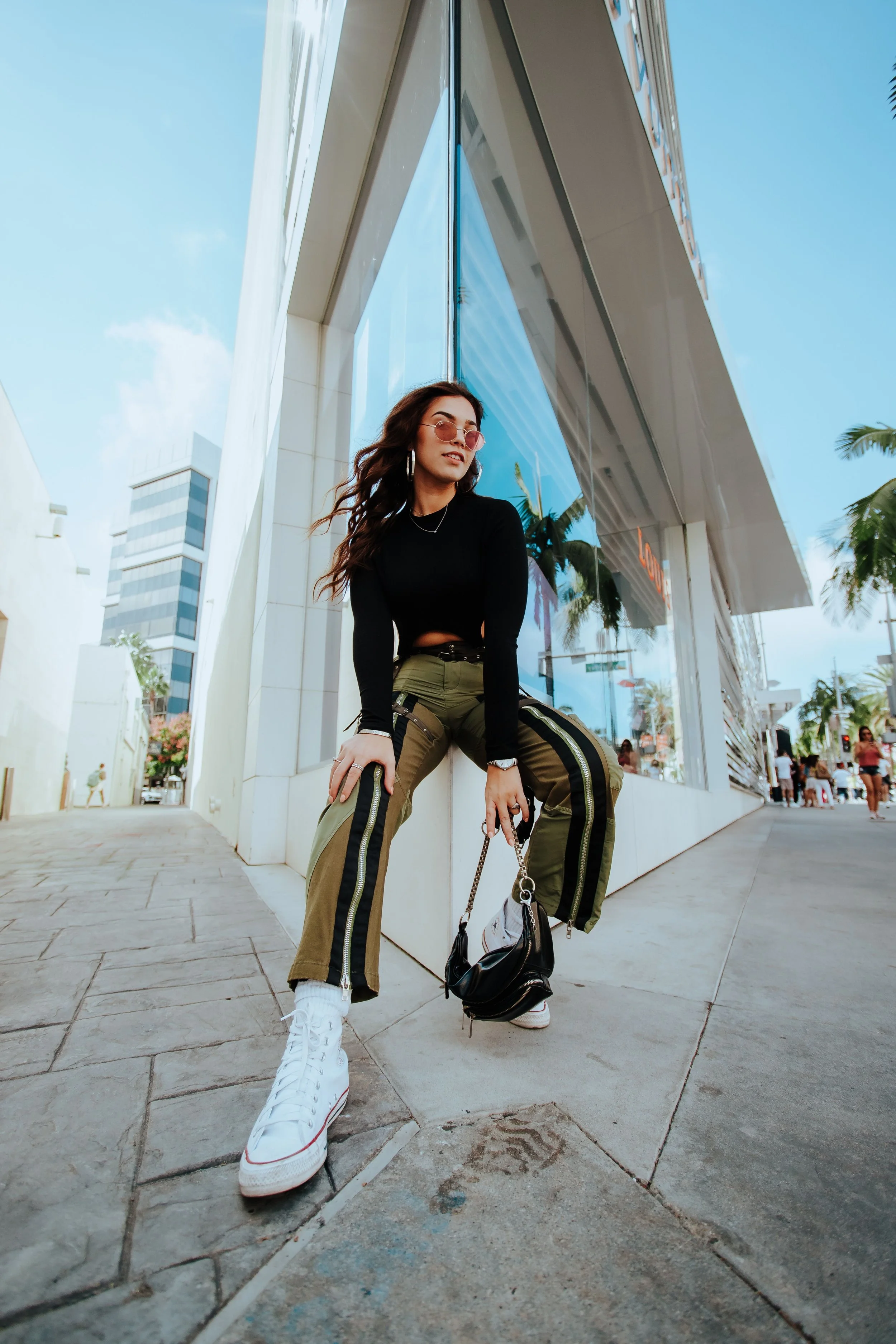 A young woman with long brown hair, wearing pink-tinted sunglasses, a black long-sleeve top, olive green cargo pants with black stripes, white sneakers, and jewelry, is sitting on a ledge outside a modern building with reflective glass and is holding a black handbag and a dog leash, with palm trees and other pedestrians in the background.