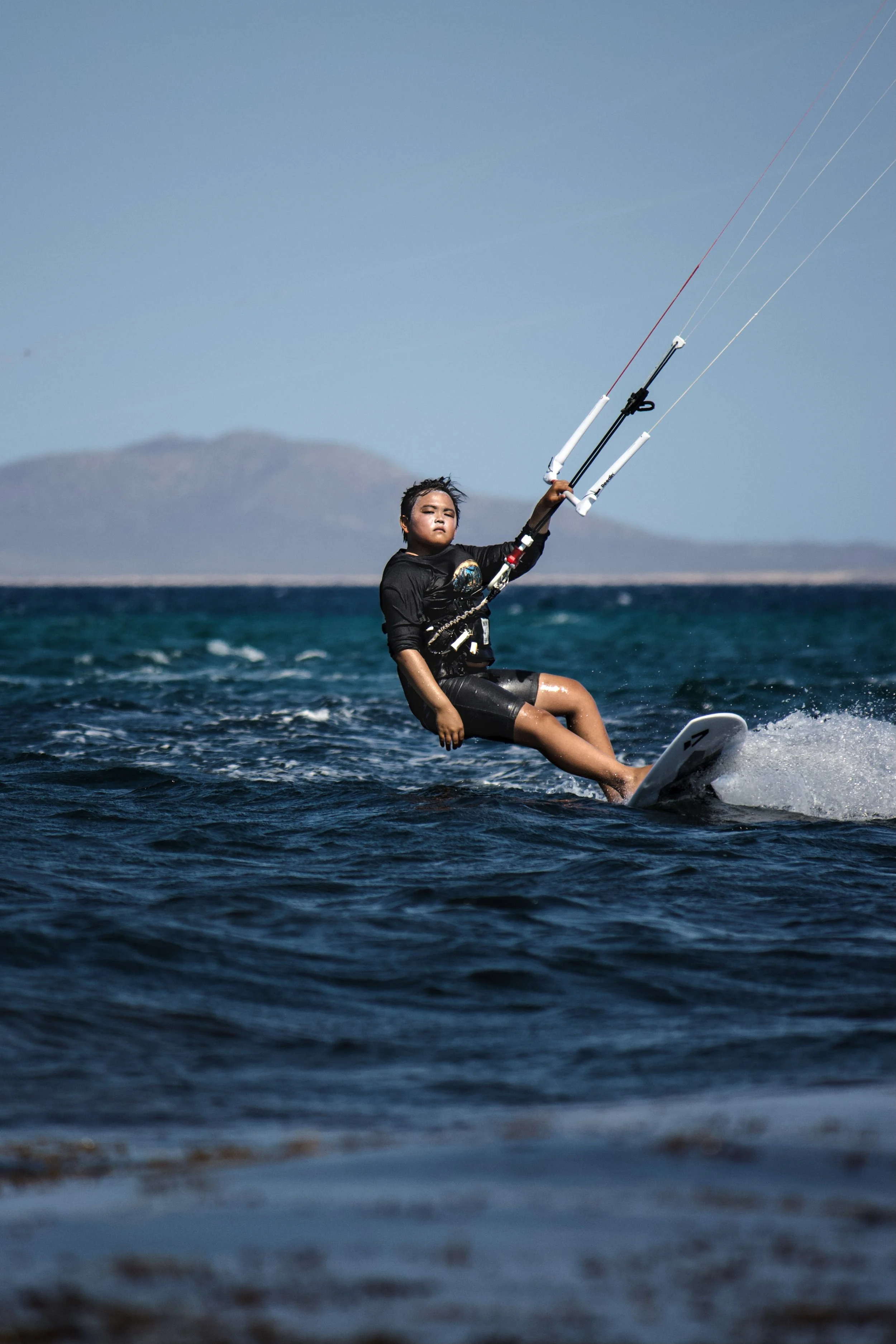 A kitesurf student sailing on a small surfboard in the water at La Ventana, with mountains visible in the background.